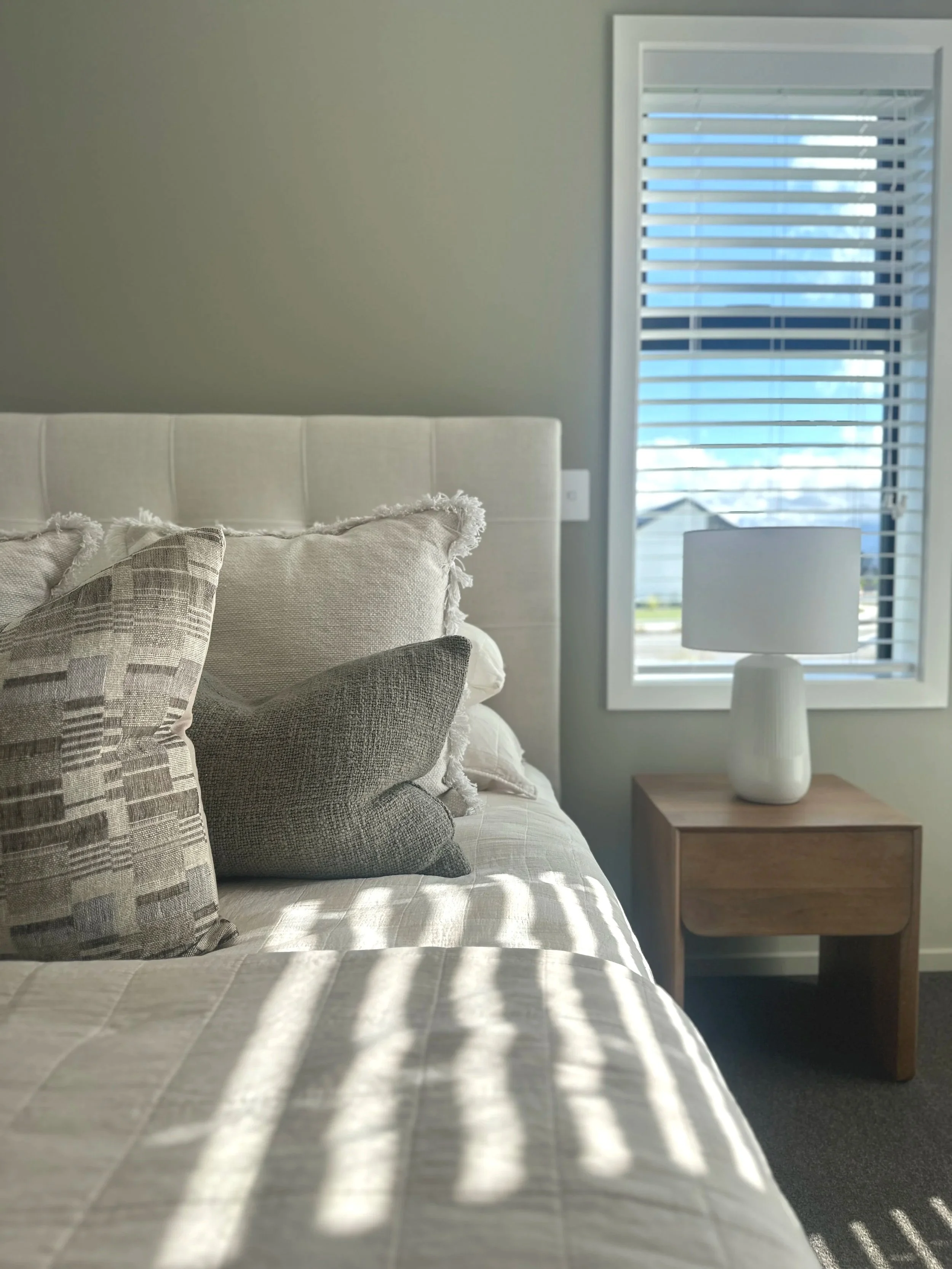 A bedroom with a bed featuring pillows, a bedside table with a white lamp, a window with blinds, and sunlight casting shadows on the bed.