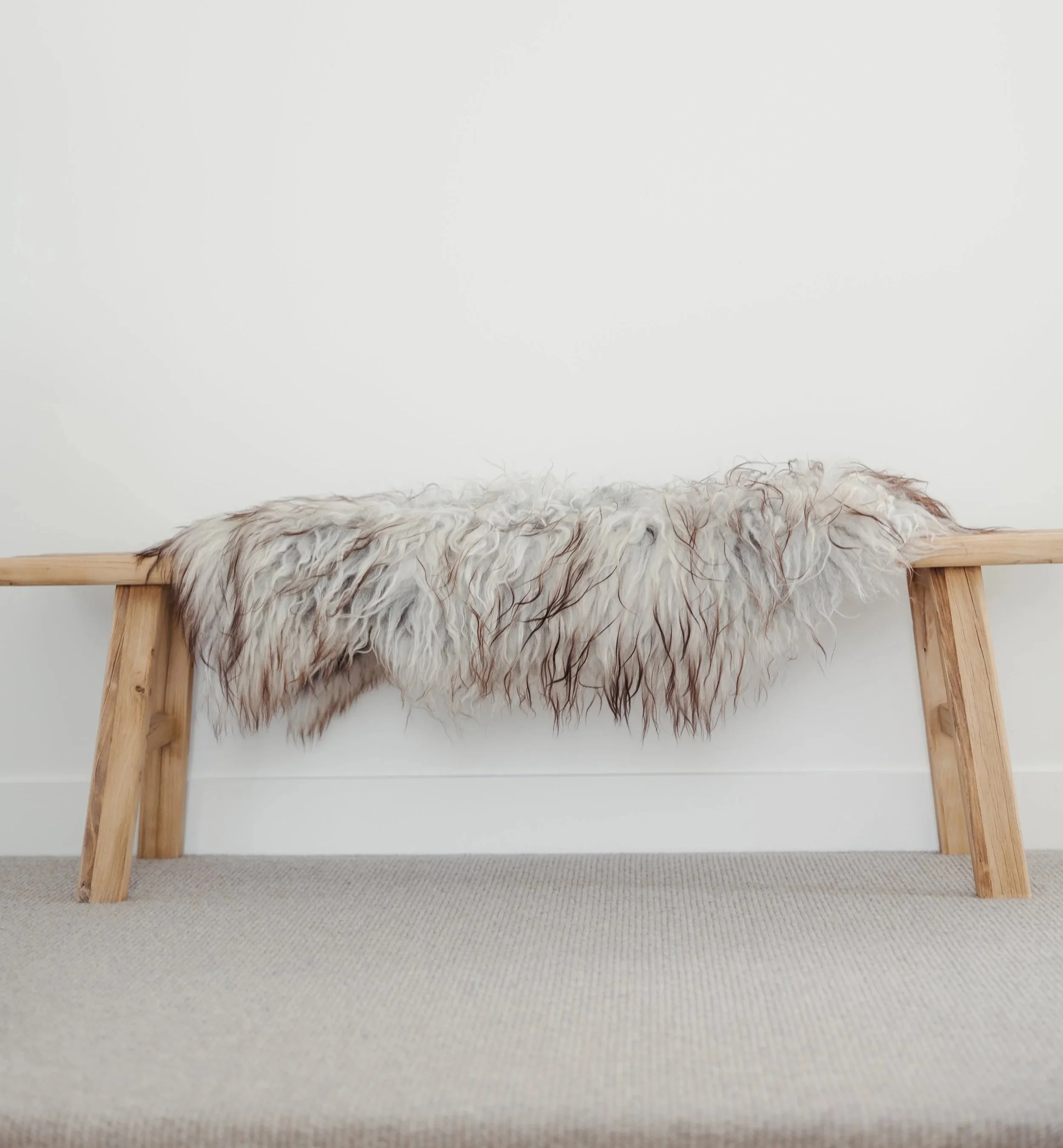 View of a wooden bench with a white, black, and brown fur rug draped over it, placed against a plain white wall and beige carpeted floor.