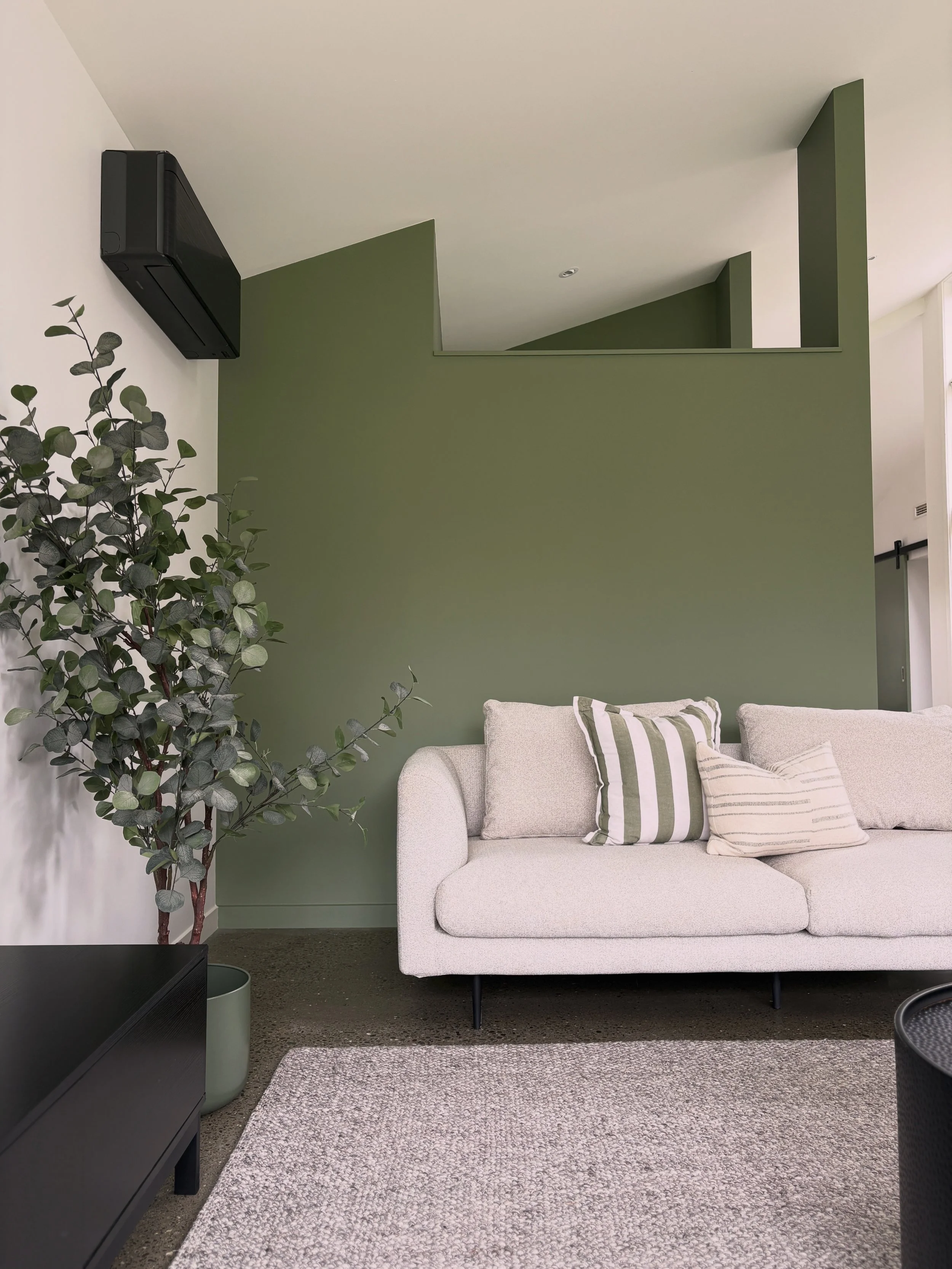 Living room corner with a white sofa, green and white striped pillows, a green accent wall, a potted eucalyptus plant, a gray textured rug, a black media console, and part of a sliding barn door.