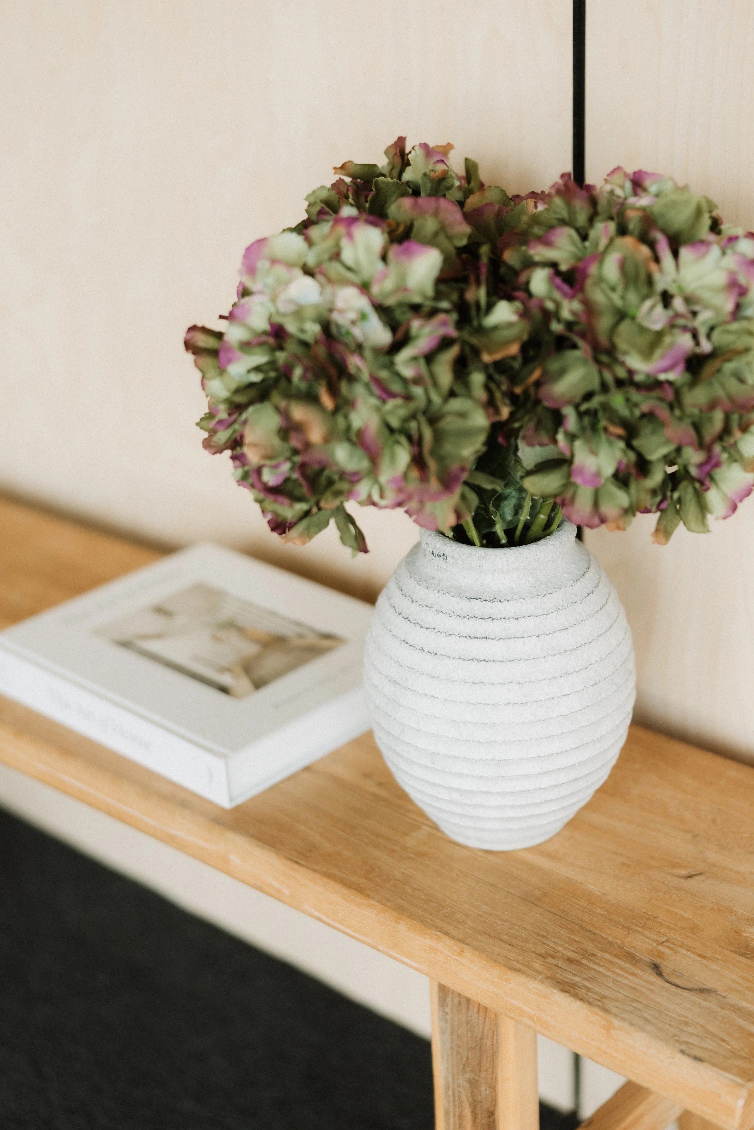A white ceramic vase with horizontal ridges, containing pink and green hydrangea flowers, sitting on a wooden table. A picture frame is slightly visible in the background.