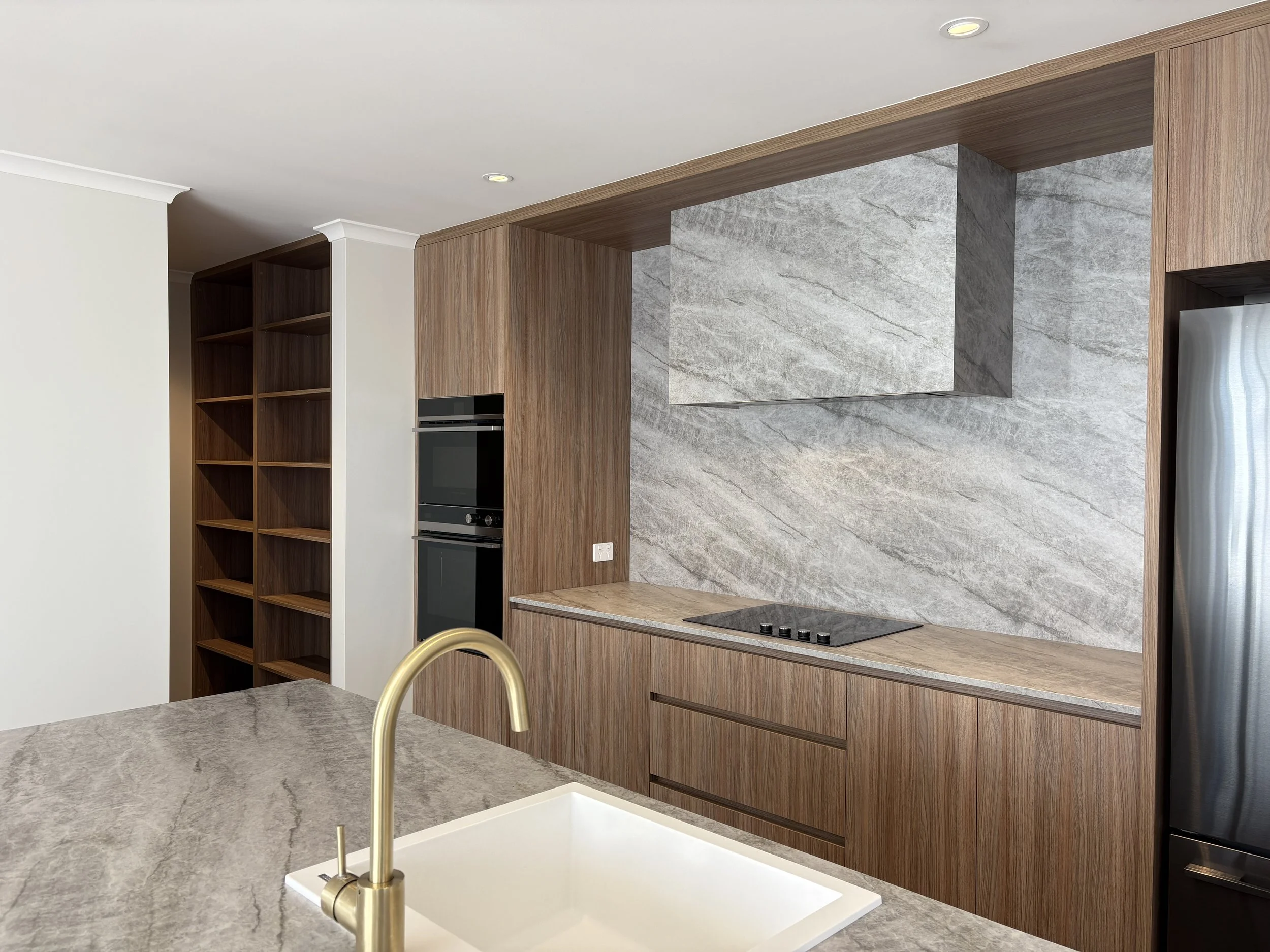 Modern kitchen with beige marble countertop, white sink with gold faucet, wooden cabinets, a black oven, and a gray marble backsplash.