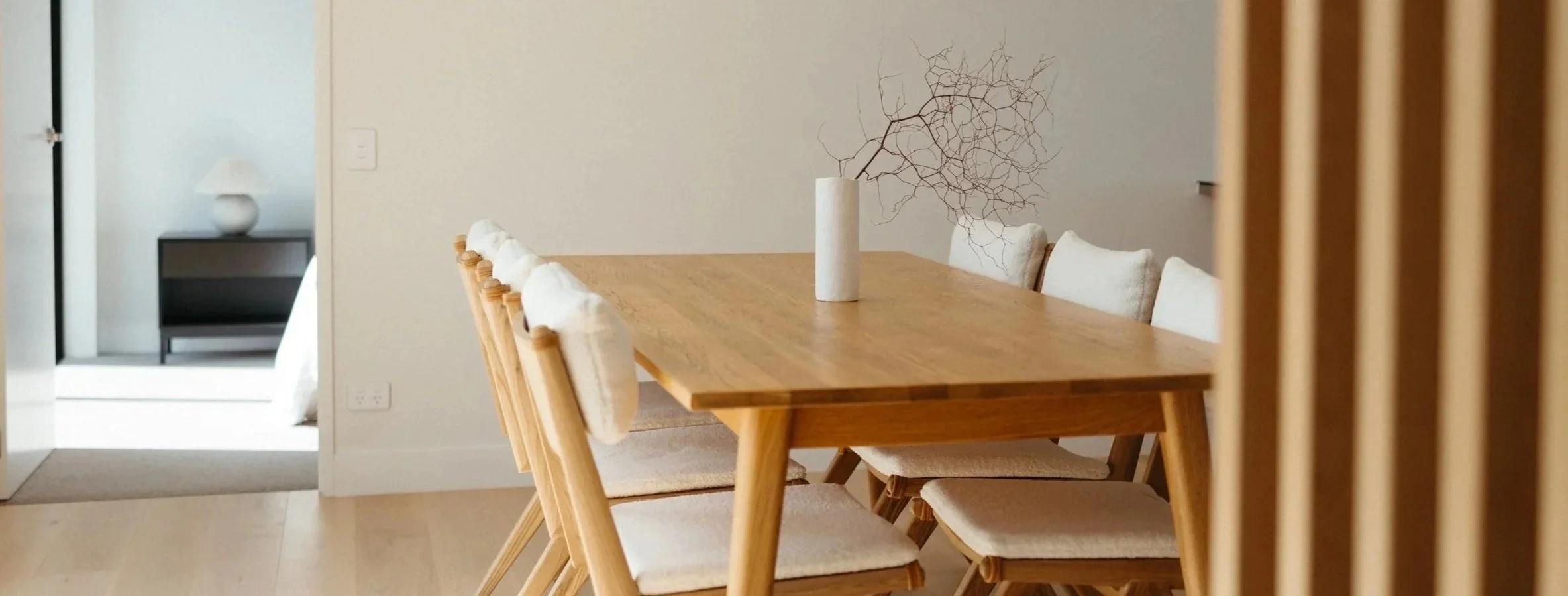 A wooden dining table with eight upholstered chairs, a white vase with decorative twigs, and a side table with a lamp in a minimalist dining room.