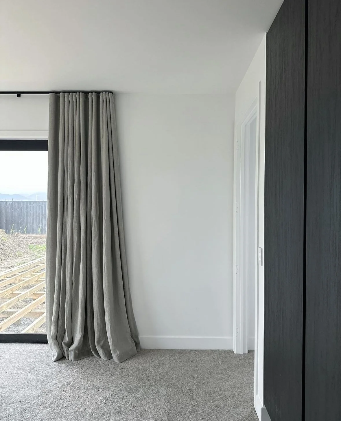 Interior of a modern room with a large sliding glass door covered with gray curtains, beige carpet, white wall, and part of a black textured wall or cabinet on the right.