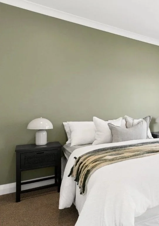 A neatly made bed with white and gray pillows, a beige and black striped blanket, a black nightstand, and a white table lamp against an olive green wall.