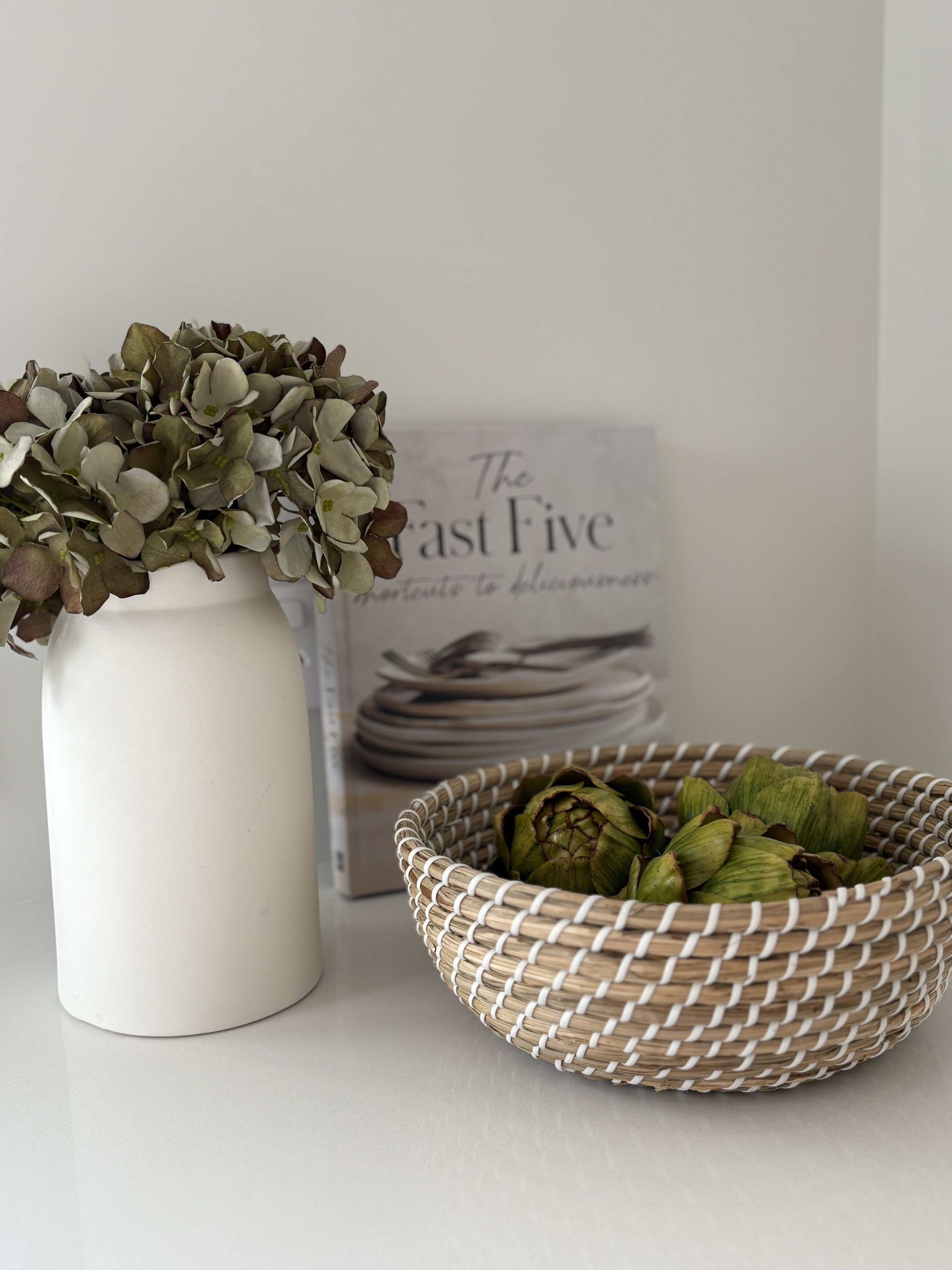 A white vase with a bunch of dried hydrangea flowers and a basket of artichokes on a white surface.
