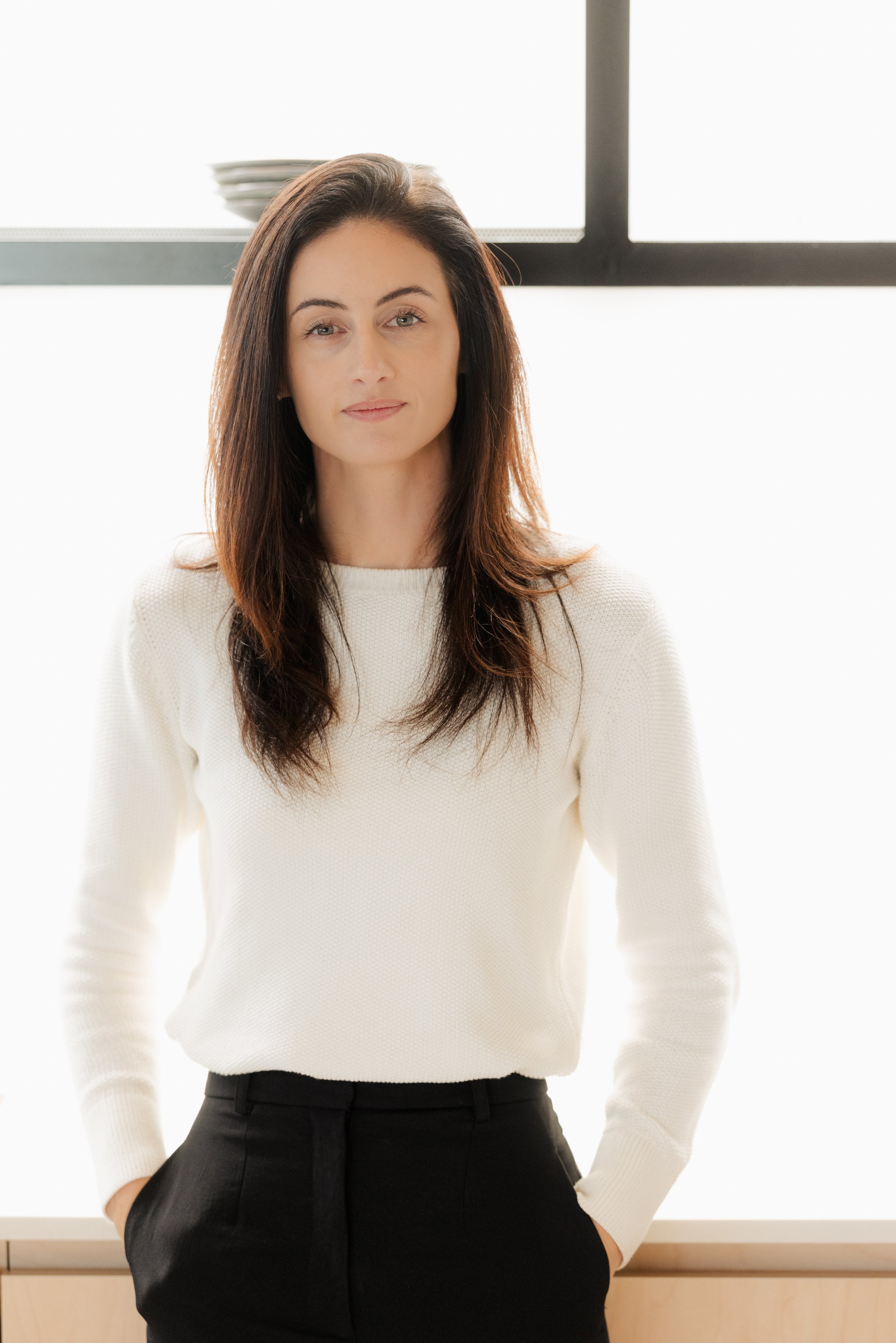 Woman with long dark hair wearing a white sweater and black pants standing indoors in front of a window with a stack of dishes on a shelf behind her.