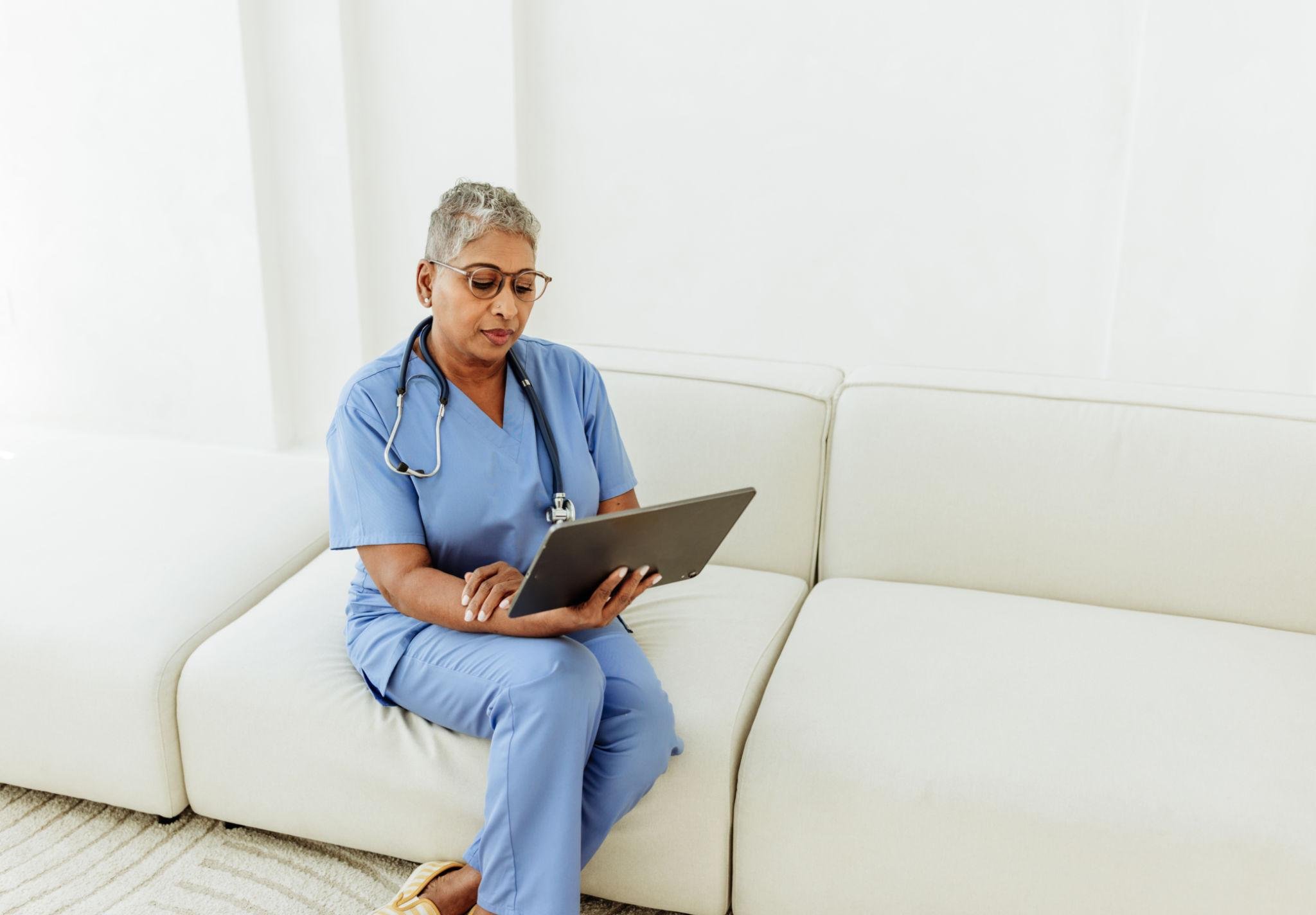 A healthcare worker in scrubs and glasses sitting on a white sofa, looking at a tablet in a bright room.