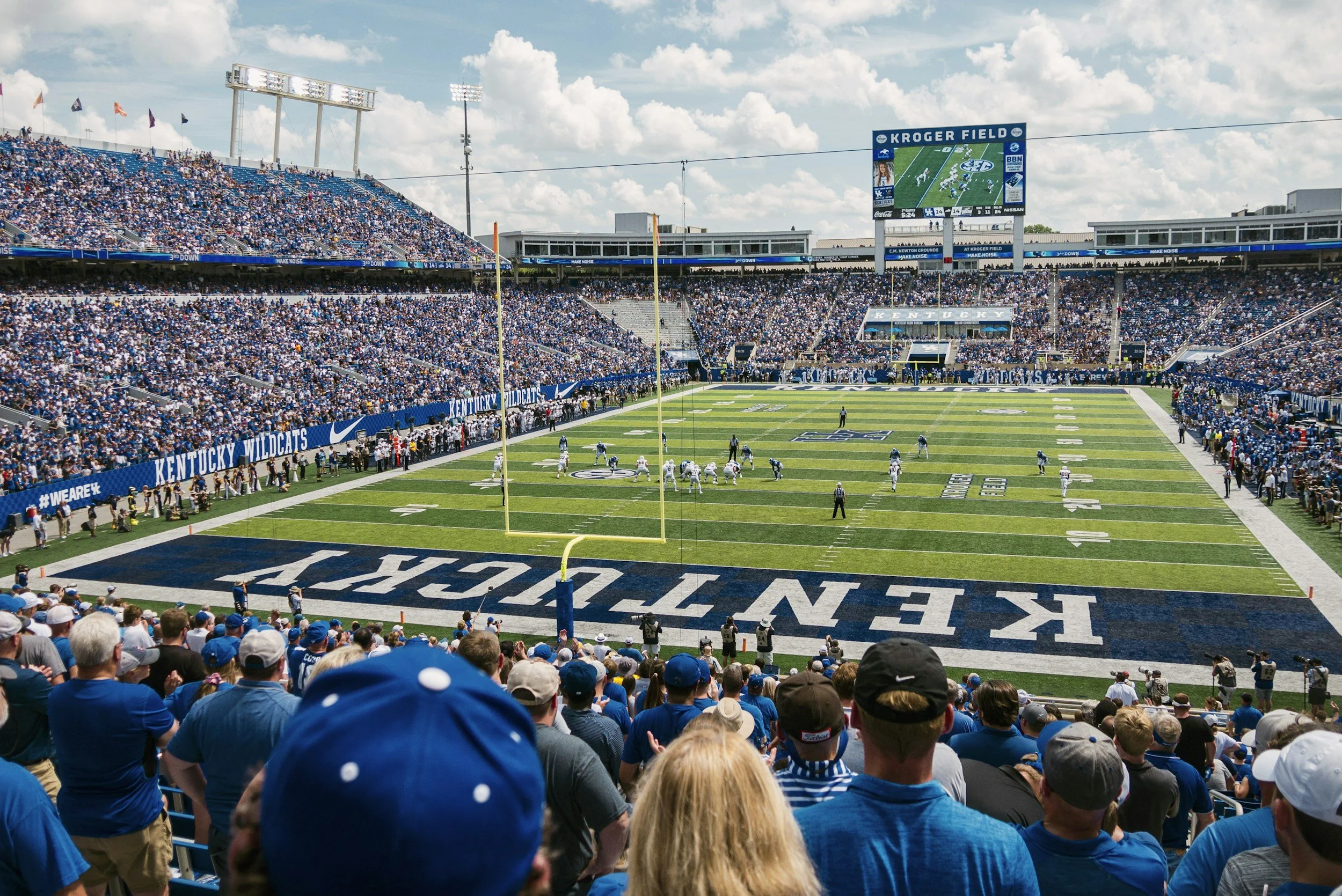 Crowd of football fans in blue shirts seated in a stadium watching a game on the field and a large screen displaying a play.