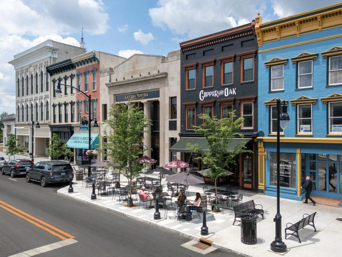 Street view of sidewalk cafes with outdoor seating and umbrellas, in front of colorful storefront buildings, and a few pedestrians walking by.