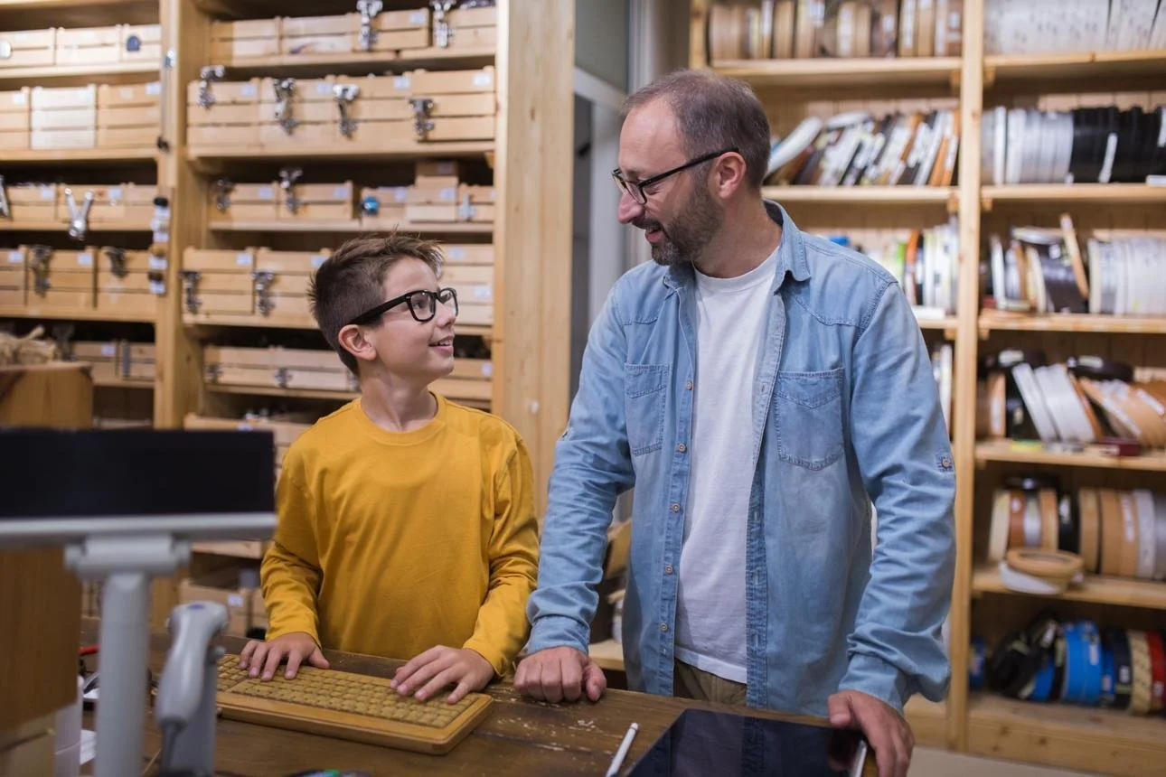 A boy with glasses talking to a man with glasses in a workshop or library with wooden shelves filled with books and tools.
