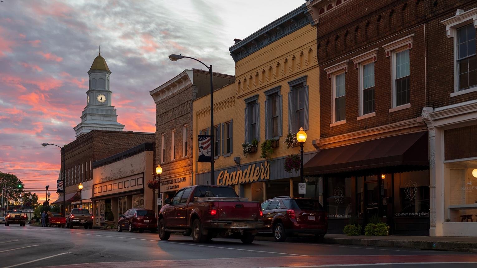 A small town street scene at sunset with historic brick buildings, parked cars, and a church with a clock tower in the background.