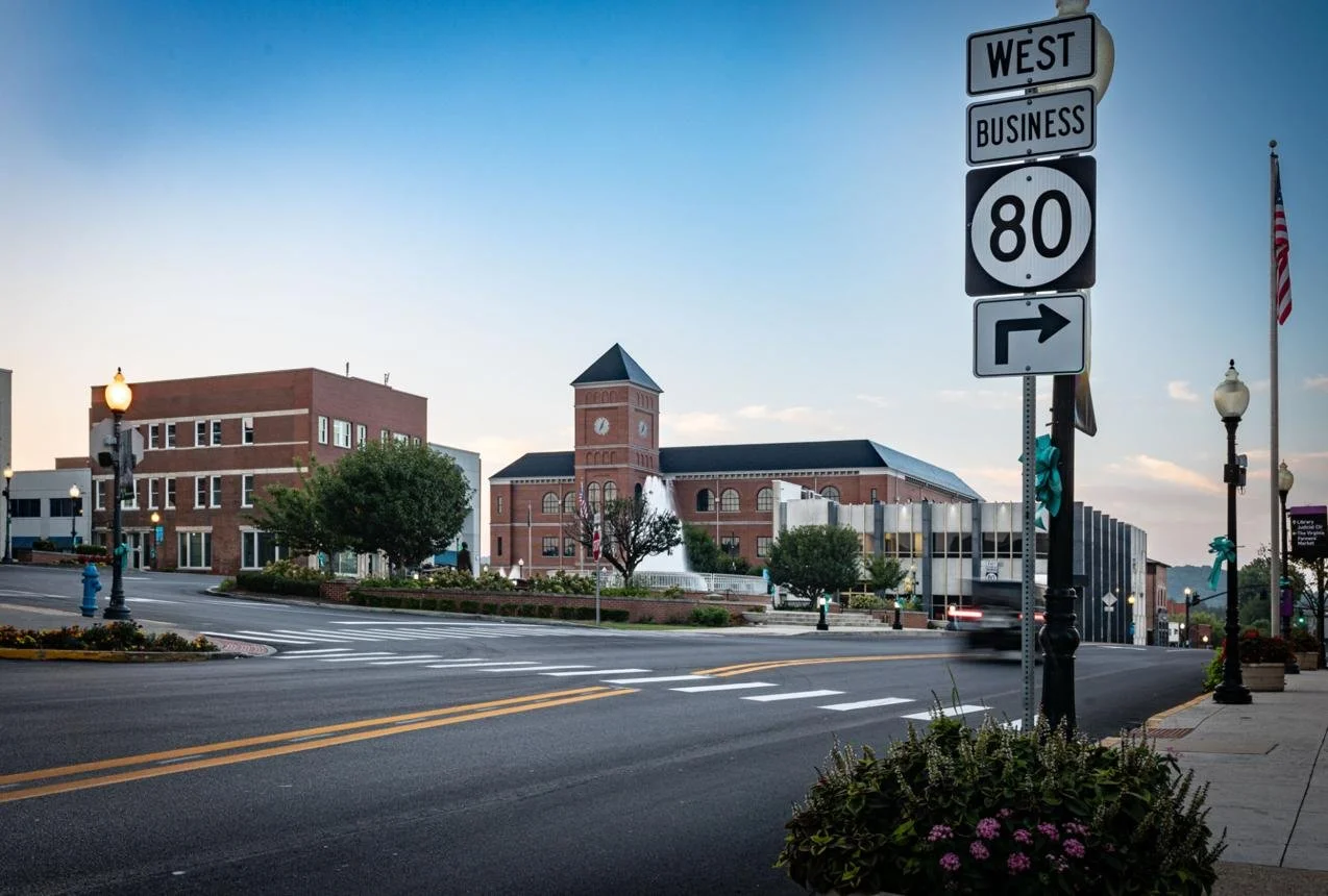 City street scene in the evening with brick buildings, a clock tower, trees, street lamps, and a traffic sign indicating westbound business route 80 with a right turn arrow. A small fountain is visible near the clock tower.