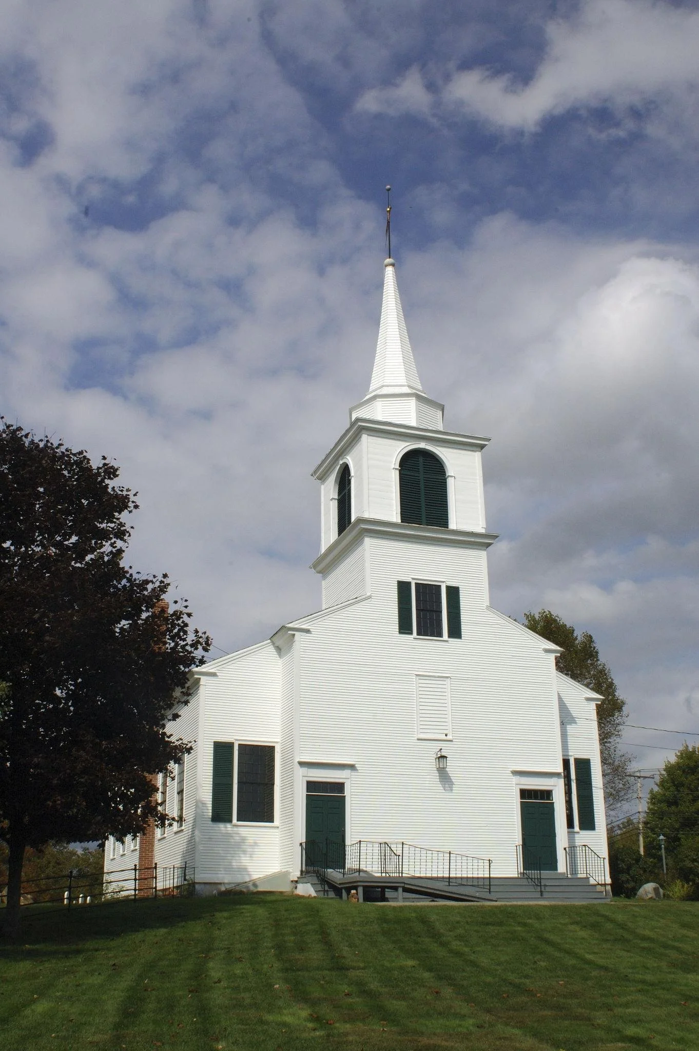 A white church with a tall steeple against a partly cloudy sky, surrounded by green grass and trees.