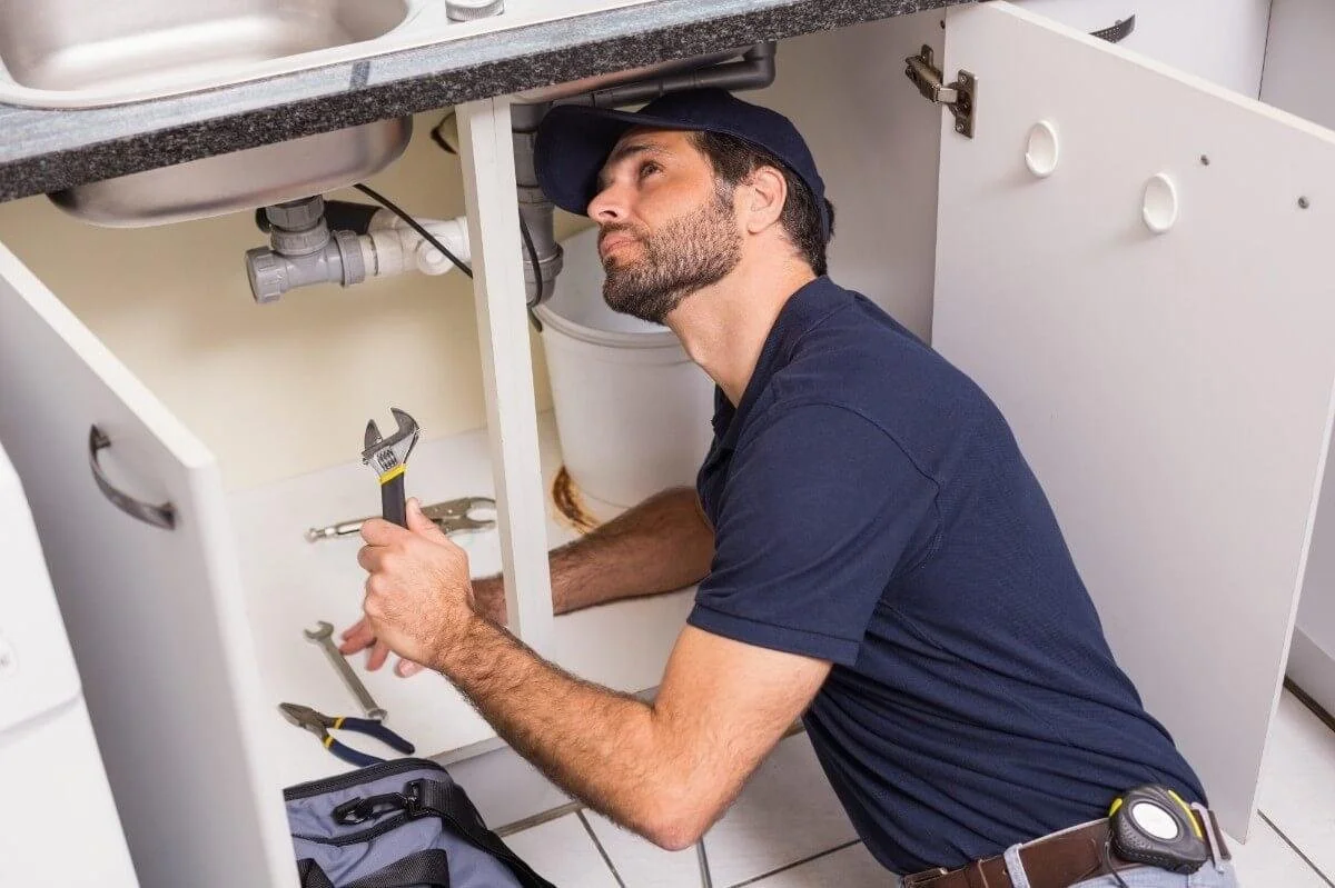 A plumber inspecting pipes underneath a kitchen sink, holding a wrench with other tools nearby.