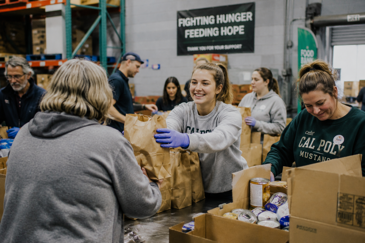 People volunteering at a food bank, packing bags of canned goods and other supplies, with a sign on the wall that says 'Fighting Hunger Feeding Hope'.
