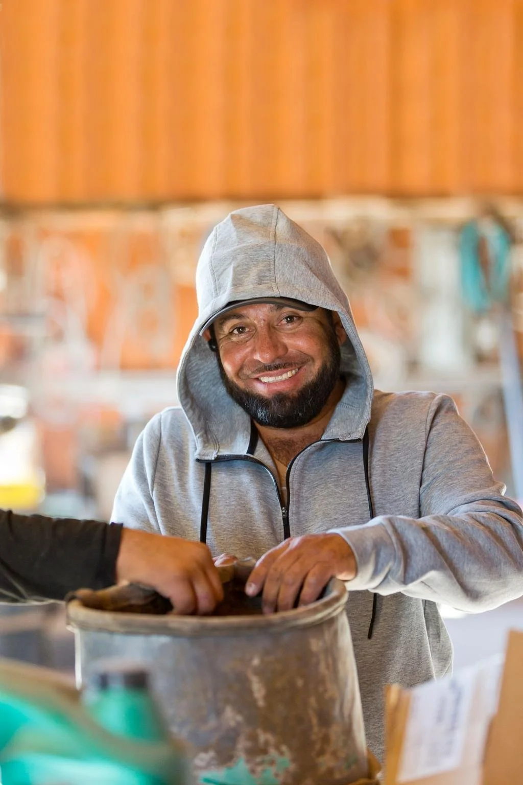 A man in a gray hoodie smiling in a workshop setting.