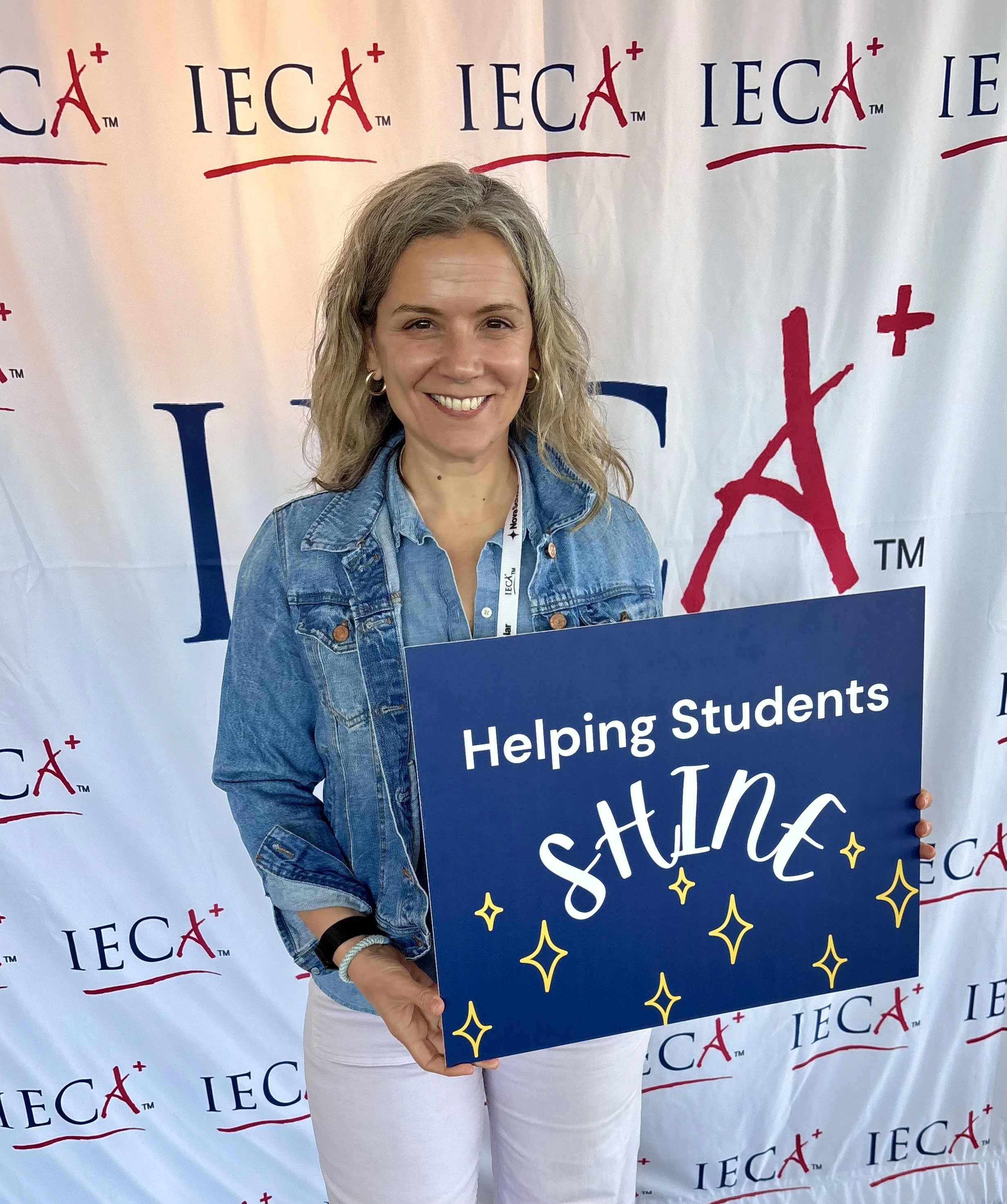 A smiling woman holding a large sign that reads "Helping Students" in front of a backdrop with the IEC and A+ logos repeatedly printed on it.