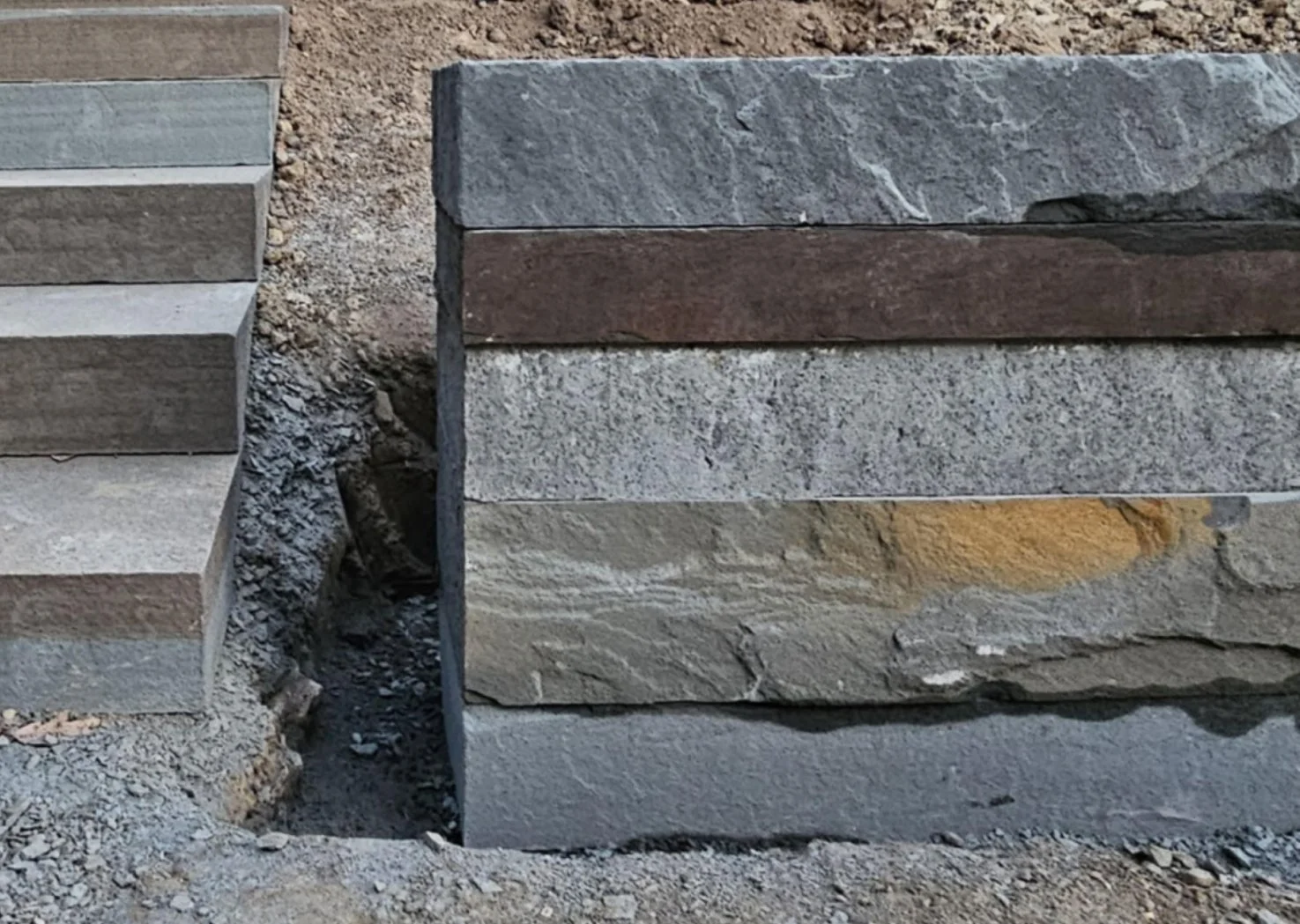 Stacked rectangular stones of varying colors and textures on a dirt ground, with some small gravel and dirt visible around them.