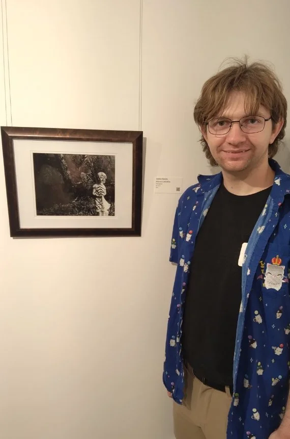 A young man wearing glasses and a blue festive jacket stands next to a framed black-and-white photograph of a child in a striped shirt in an art gallery.