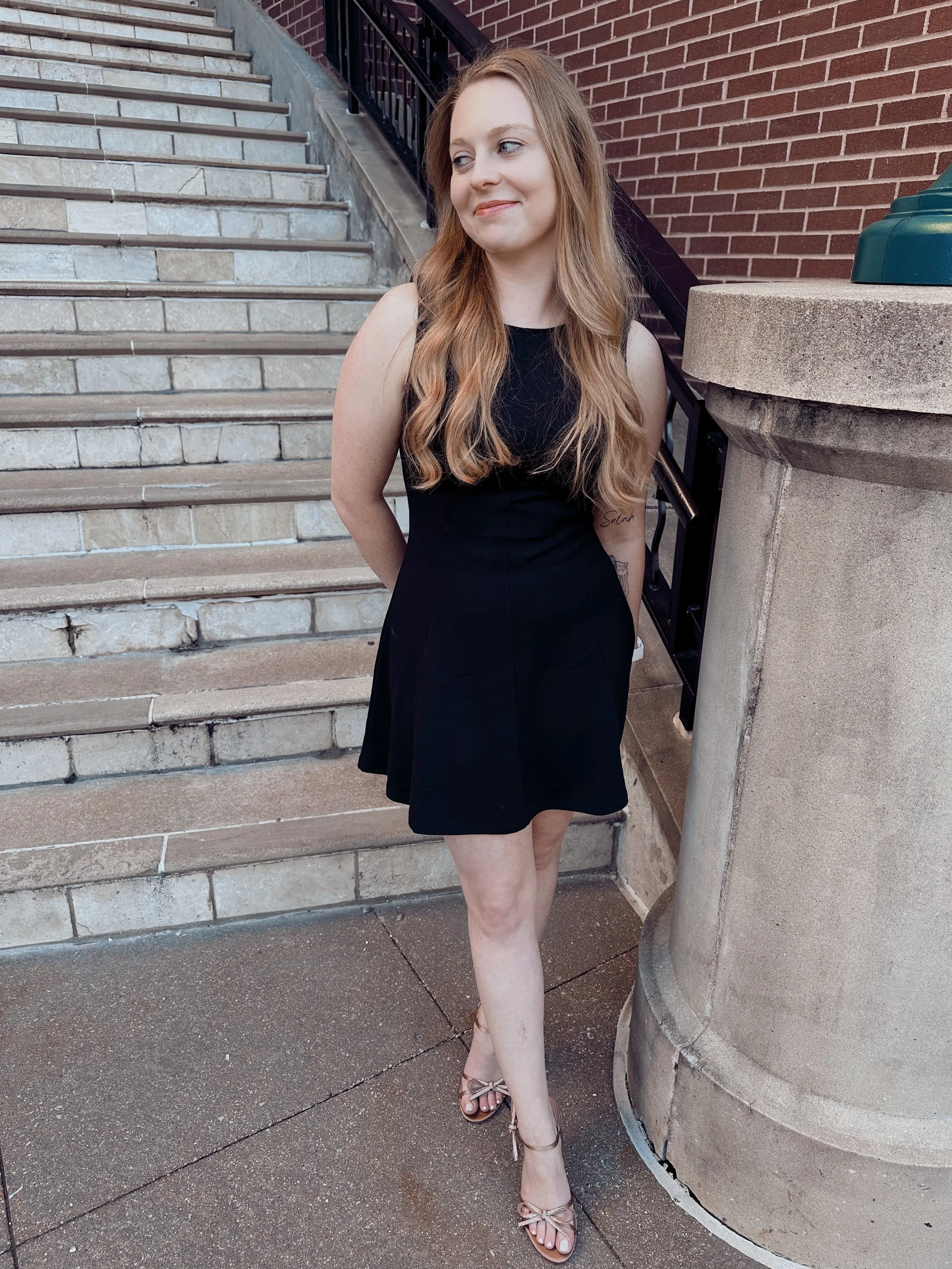 A young woman with long wavy hair wearing a black dress and sandals standing on a sidewalk near stairs and a brick building.