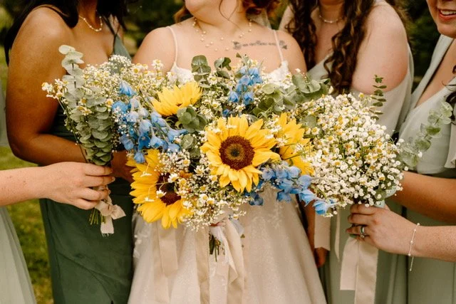 A group of women holding a large bouquet of yellow sunflowers, white baby's breath, and blue delphinium, with greenery in the background.