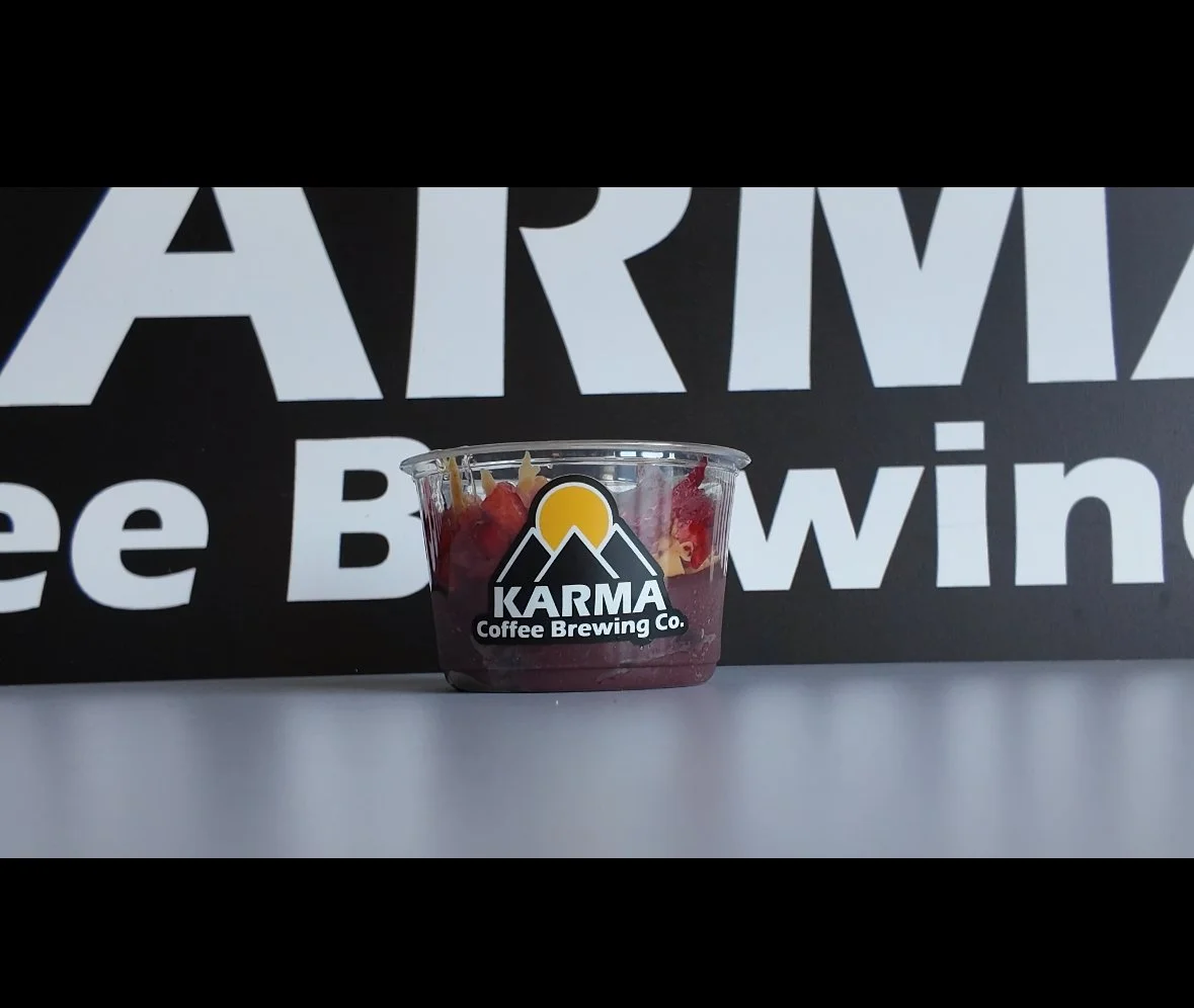 Small plastic container with a Karma Coffee Brewing Co. logo, containing a layered dessert topped with fruit, set against a black background with large white text.