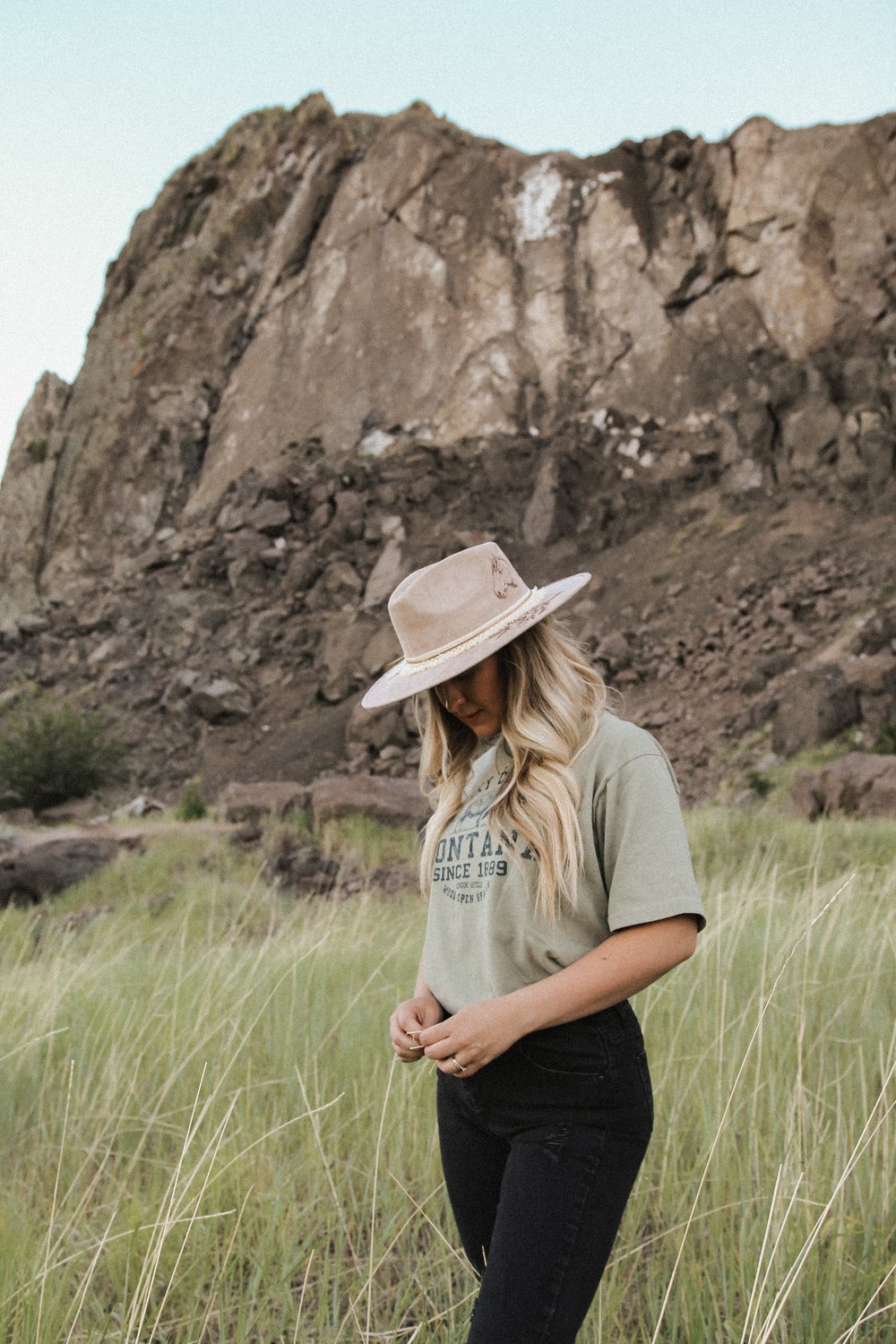 A woman wearing a beige hat and a green t-shirt stands in a grassy field with a rocky mountain in the background.