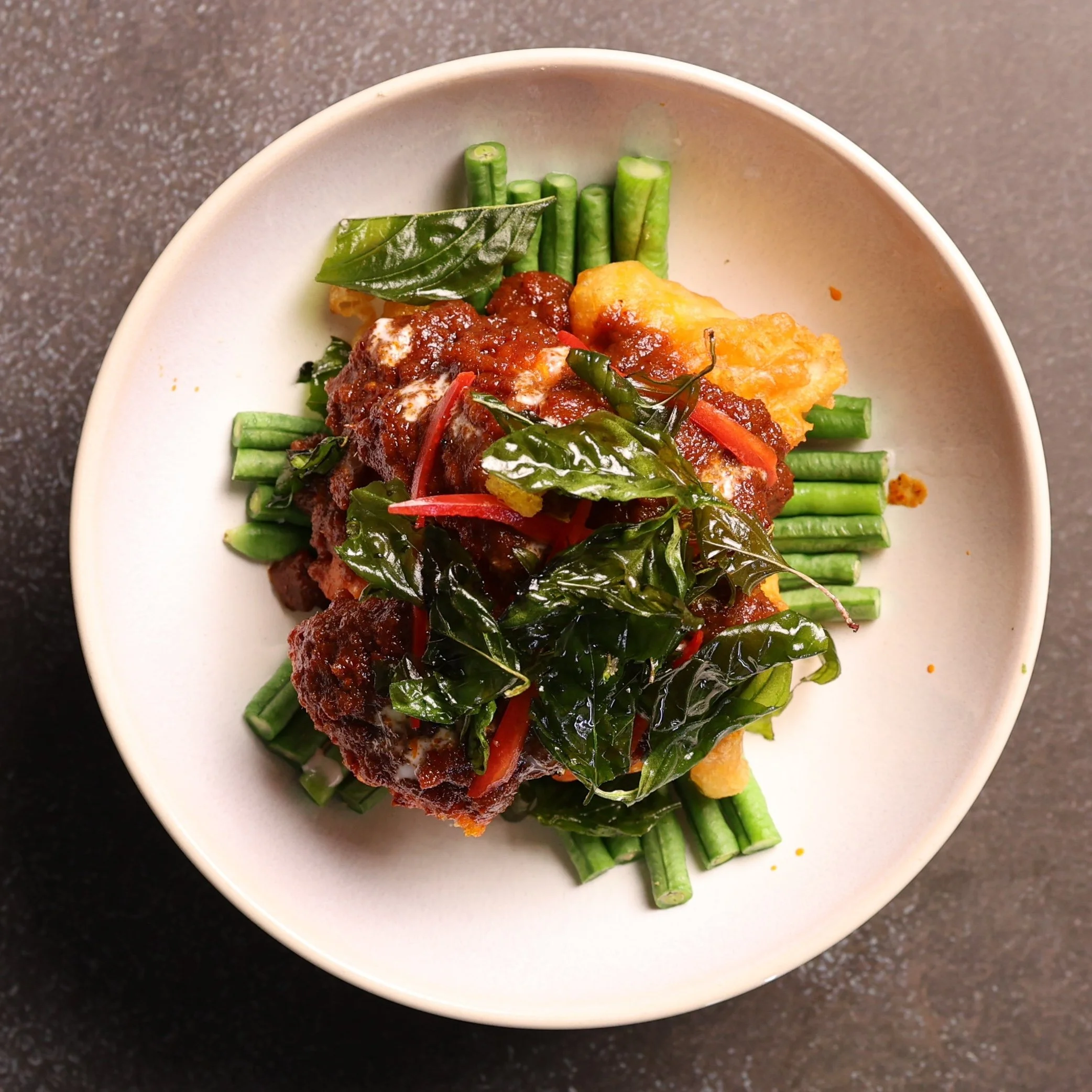 A white bowl filled with a dish consisting of fried fish topped with tomato-based sauce, served on a bed of green beans and garnished with leafy greens and chili peppers.