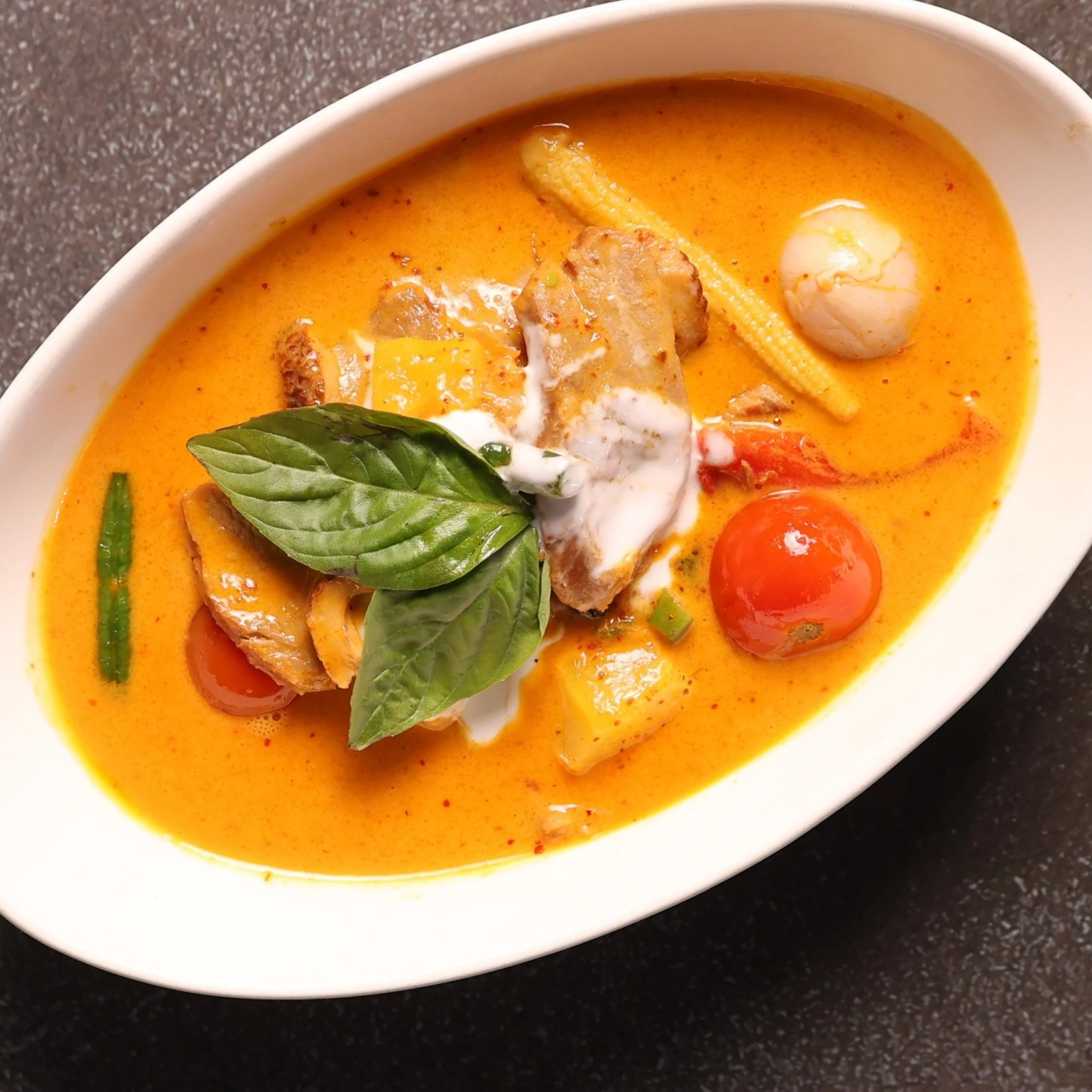 A bowl of Thai red curry with vegetables, chicken, cherry tomatoes, baby corn, and basil garnish.