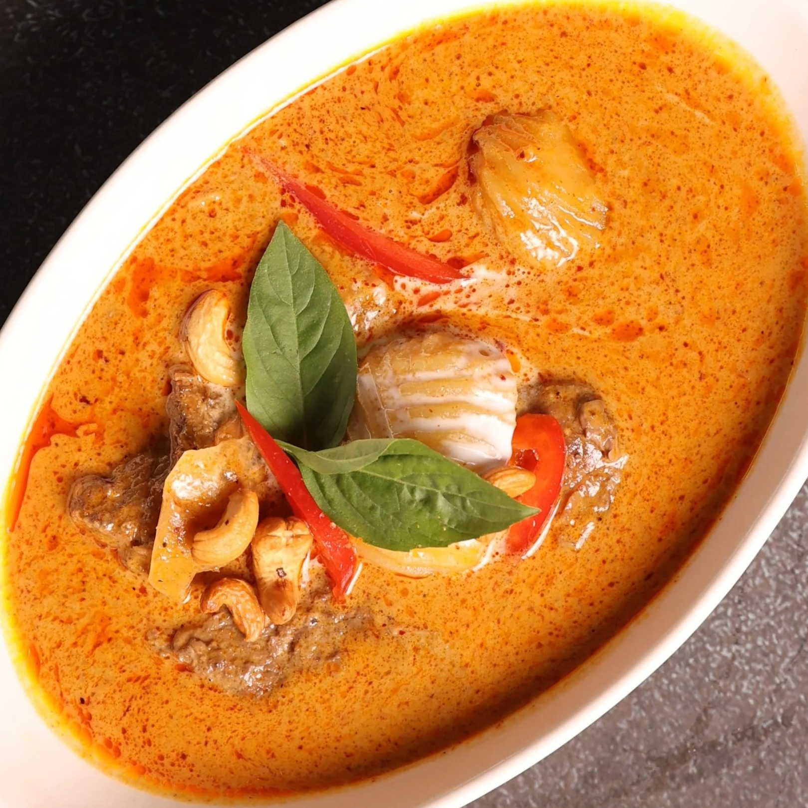 Close-up of a bowl of creamy red Thai curry with basil leaves, red chili pepper slices, cashews, and pieces of chicken or seafood.