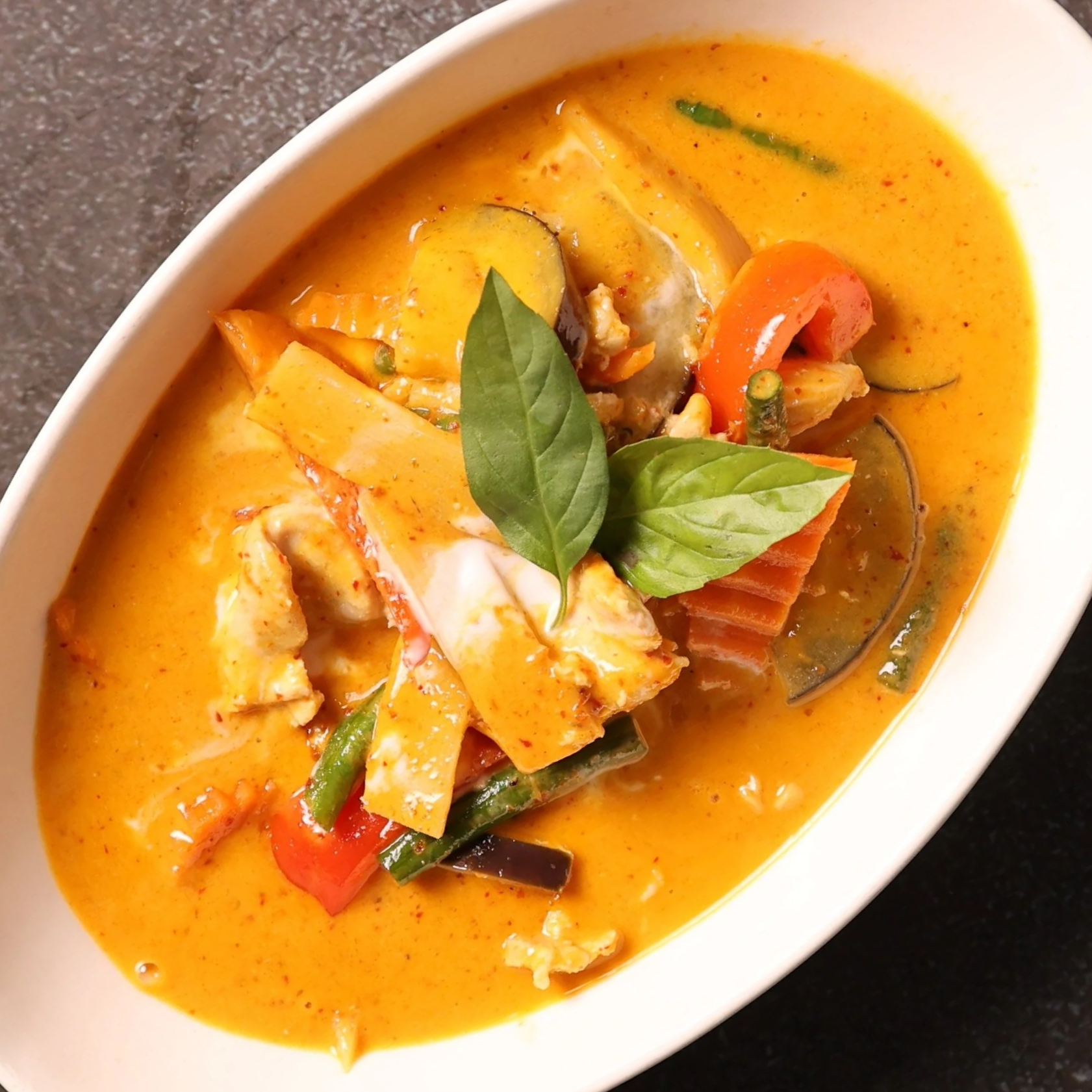 Bowl of vegetable curry garnished with fresh basil leaves, featuring carrots, zucchini, bell peppers, and eggplant in a creamy, orange-colored sauce.