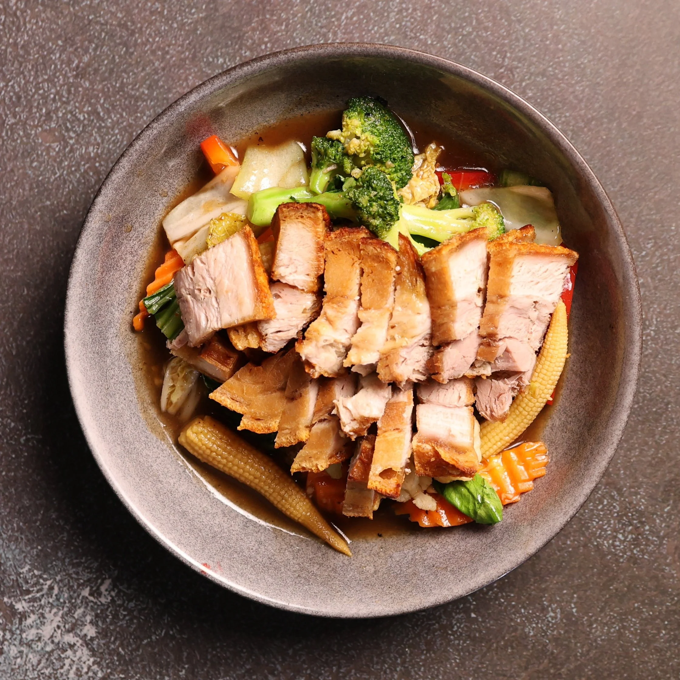 A bowl of Asian vegetable soup with chopped broccoli, carrots, cabbage, baby corn, and slices of roasted pork.
