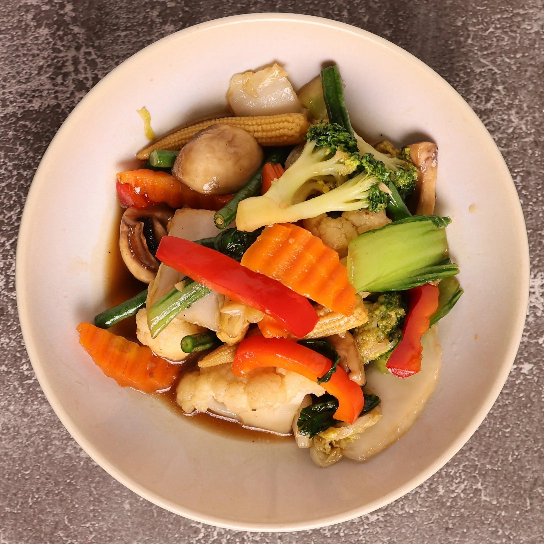 A white bowl filled with a variety of cooked vegetables including broccoli, cauliflower, red and orange bell peppers, baby corn, zucchini, mushrooms, and carrots, placed on a speckled gray countertop.