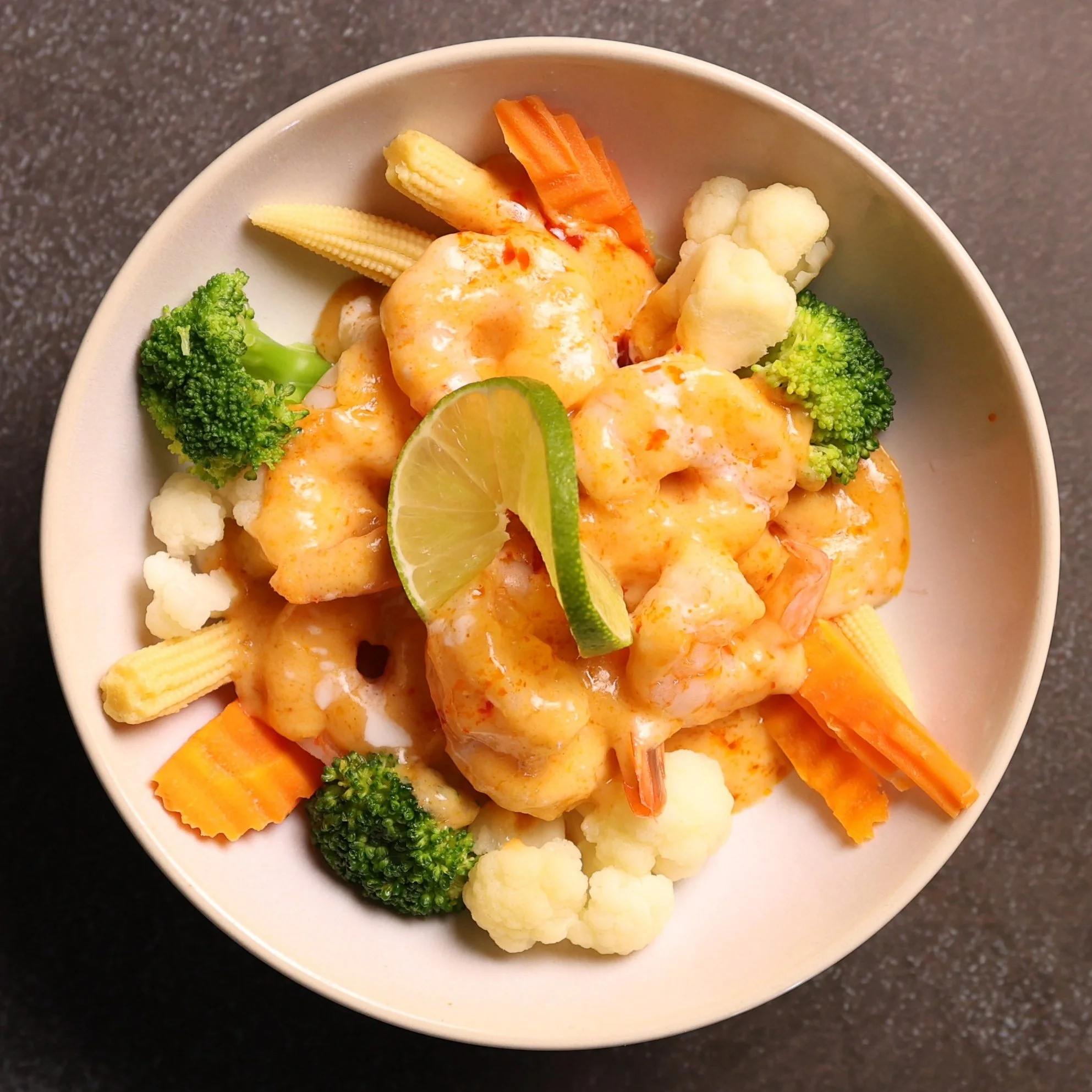 A bowl filled with cooked vegetables including broccoli, cauliflower, baby corn, carrots, and lime wedges, with a creamy orange sauce drizzled over the vegetables.