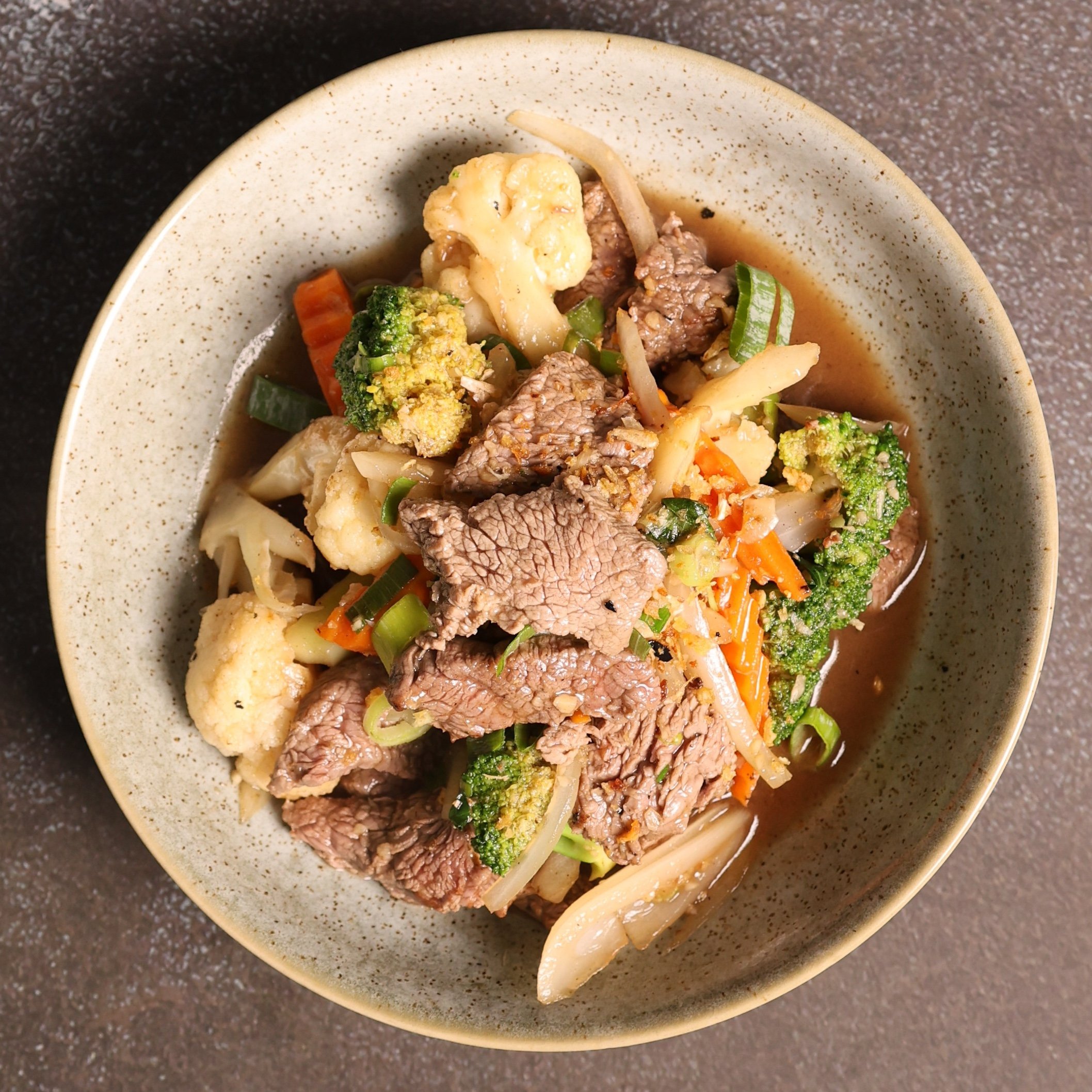 A beige bowl with speckles filled with beef and vegetable soup, including broccoli, cauliflower, carrots, and green onions on a dark speckled countertop.