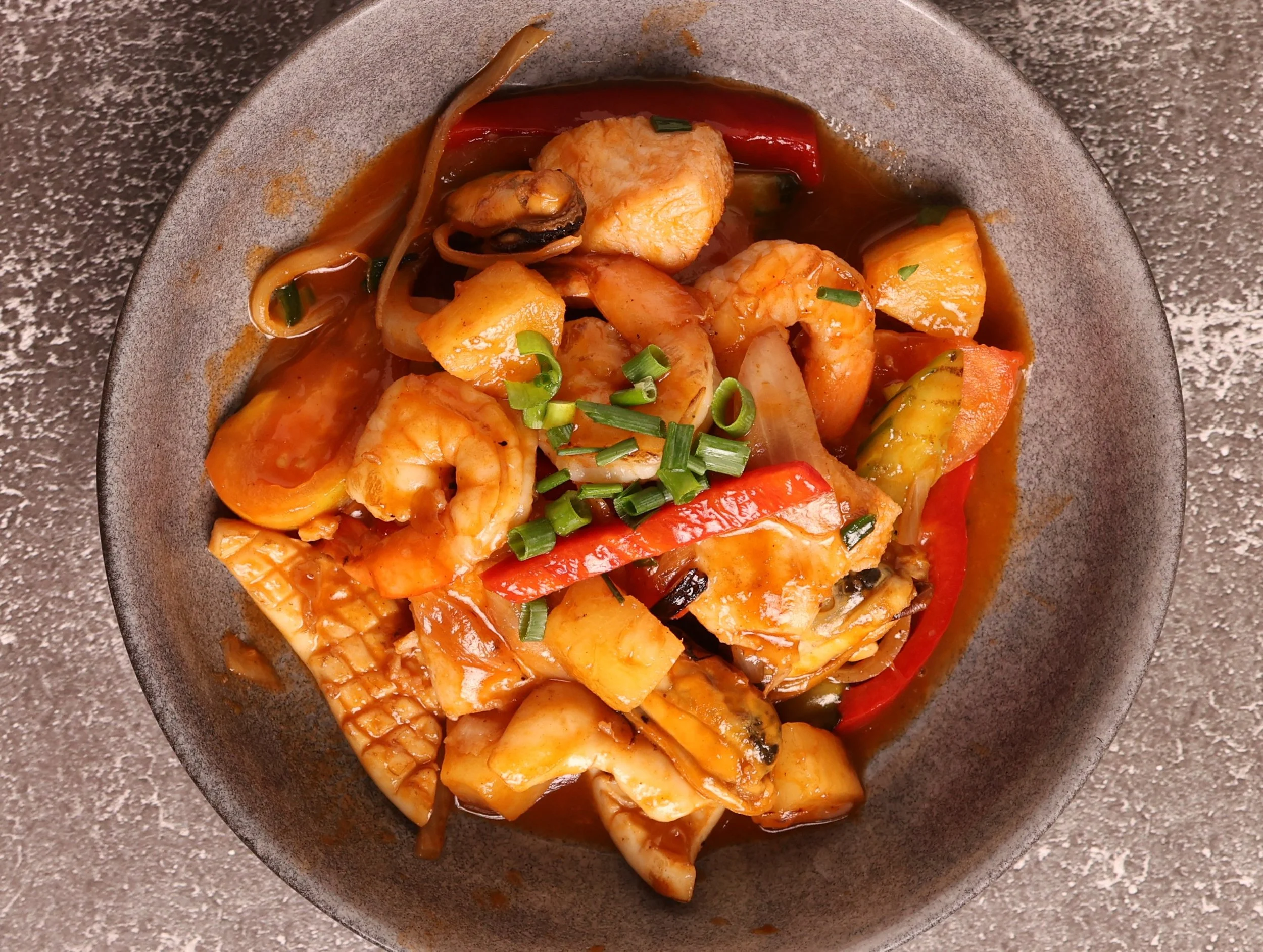 A bowl of seafood stew with shrimp, fish, squid, vegetables, and garnished with chopped green onions.