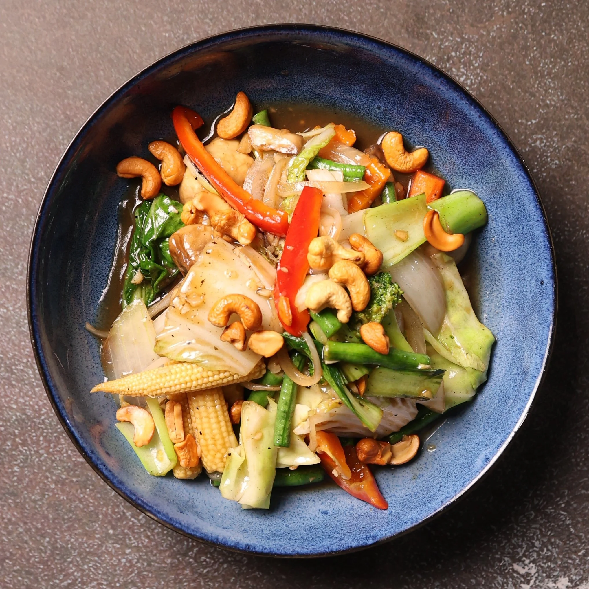 A blue bowl containing a stir-fry of vegetables including red bell peppers, green beans, broccoli, carrots, baby corn, onions, and cashew nuts, placed on a textured surface.