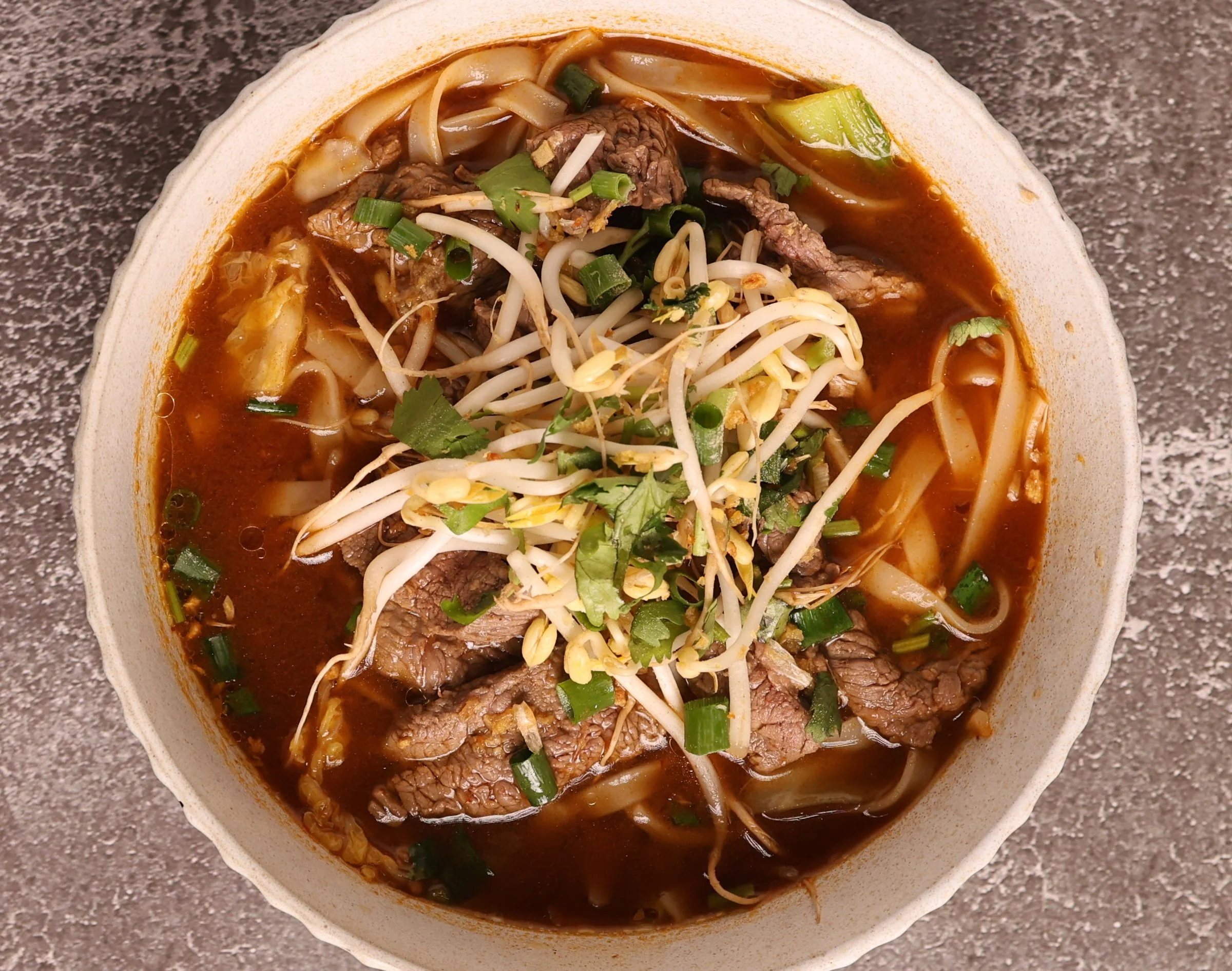 A bowl of beef pho with rice noodles, sliced beef, bean sprouts, chopped green onions, and herbs, with a dark broth on a gray textured surface.