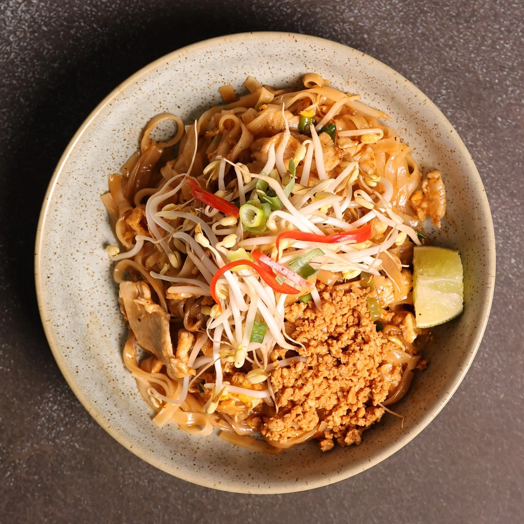 Close-up of a bowl of Asian stir-fried noodles with bean sprouts, chopped green onions, red pepper slices, ground meat, and a lemon wedge on a dark speckled countertop.