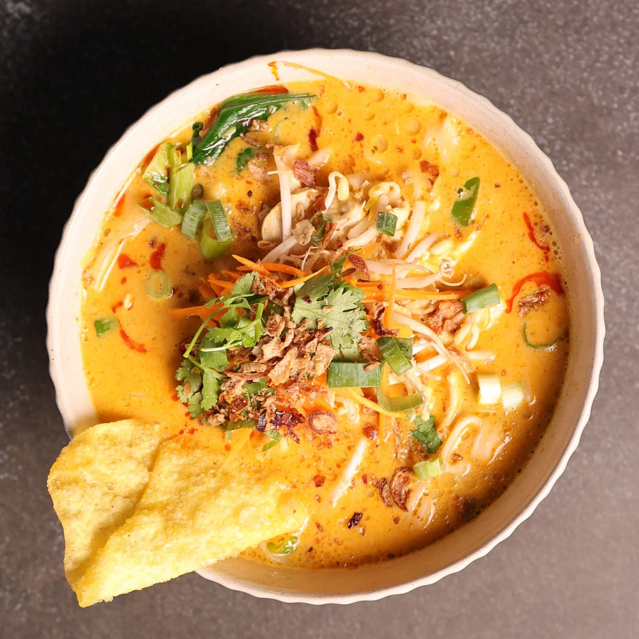 Close-up of a bowl of spicy Vietnamese noodle soup with toppings of chopped green onions, cilantro, bean sprouts, shredded carrots, and crispy fried shallots, served on a dark surface.
