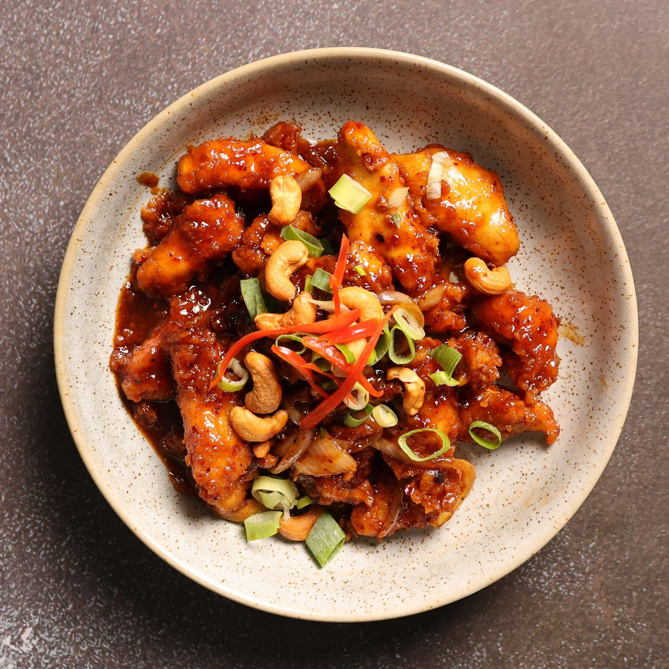 A bowl with an Asian stir-fry dish containing chicken pieces in a dark sauce, garnished with chopped green onions, red bell peppers, and cashews, on a dark speckled surface.
