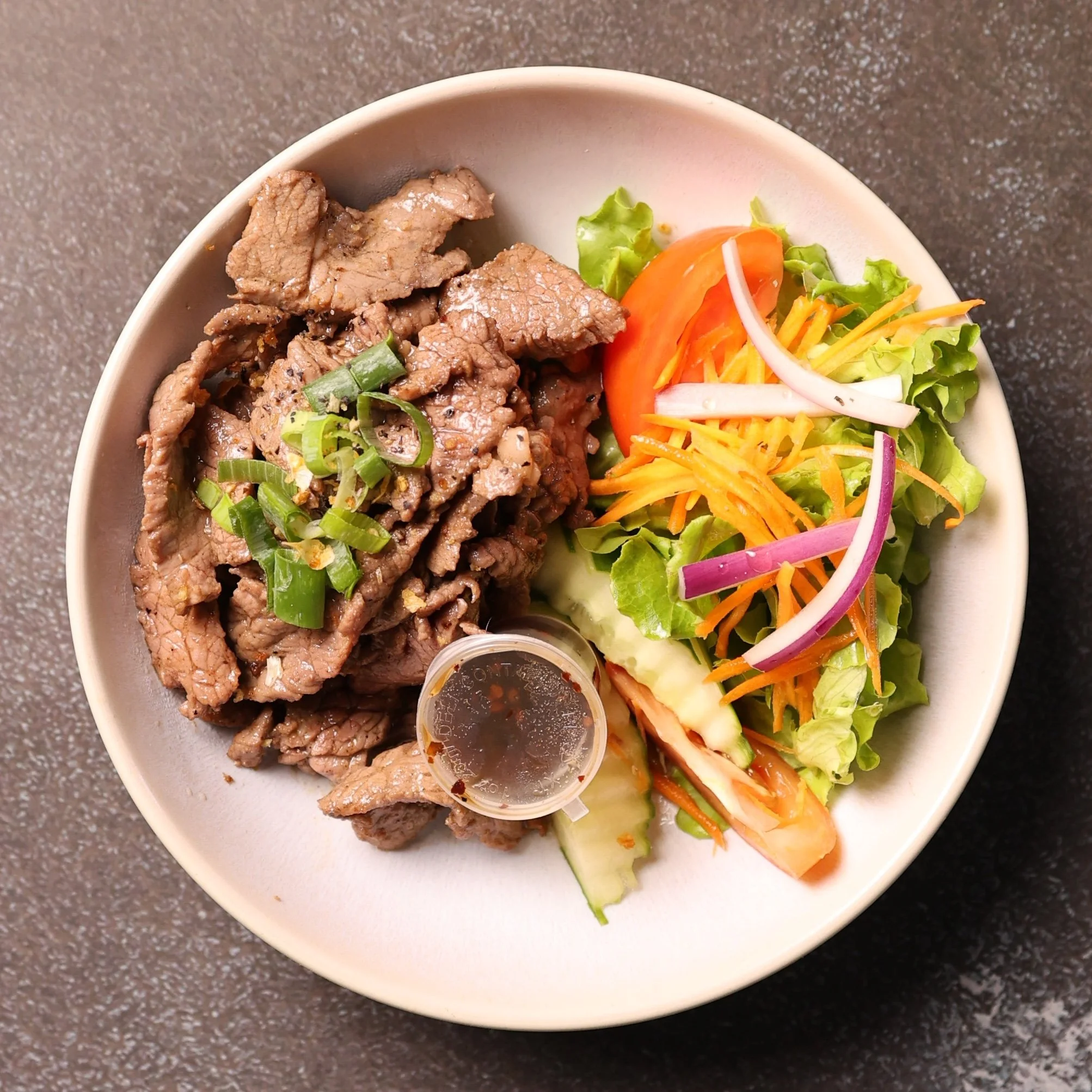A bowl of food containing cooked beef strips with chopped green onions, a small cup of dipping sauce, and a side salad with lettuce, tomato, shredded cheese, carrot, radish, and red onion.