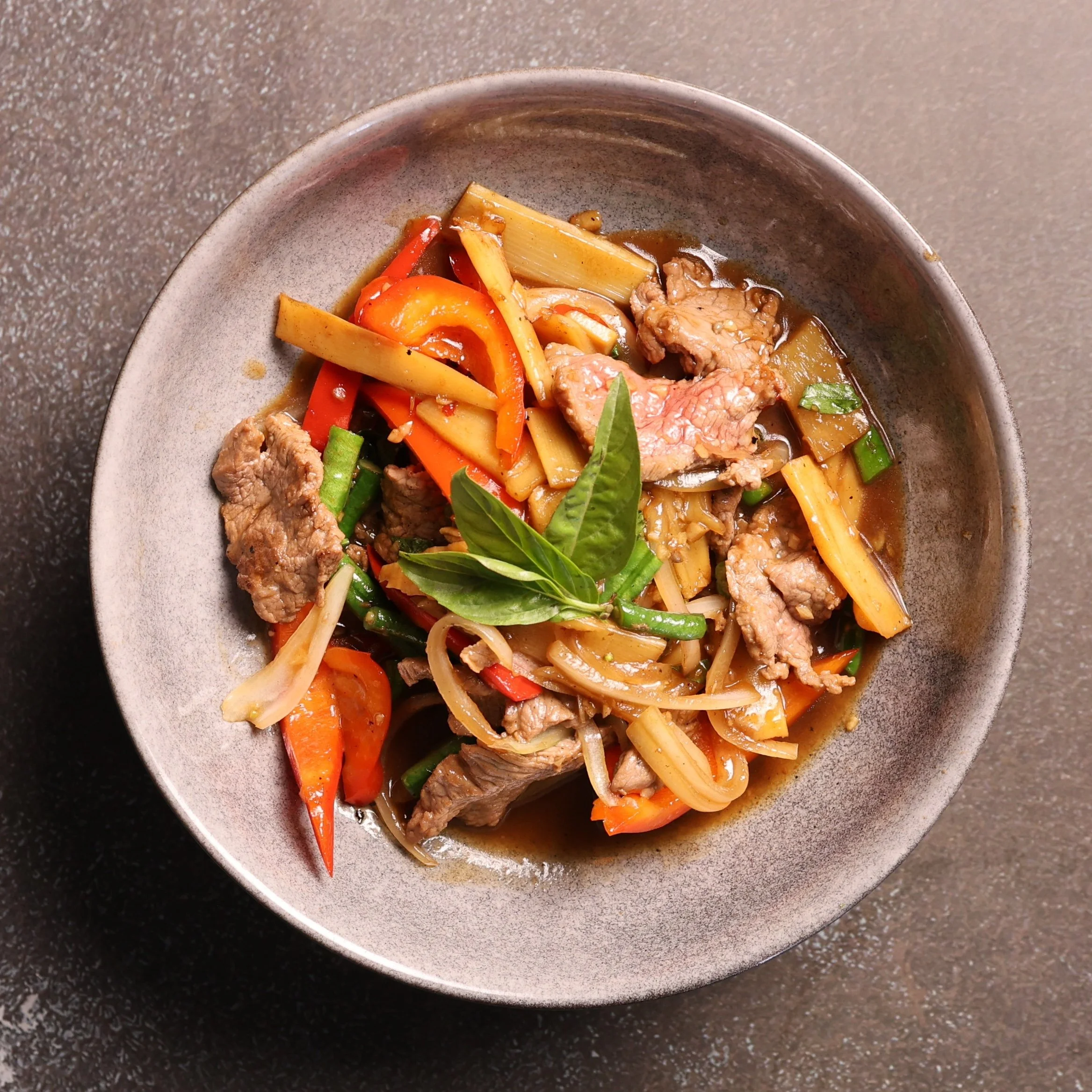 A bowl of Asian-style noodle soup with sliced beef, carrots, bell peppers, green onions, and herbs in broth.
