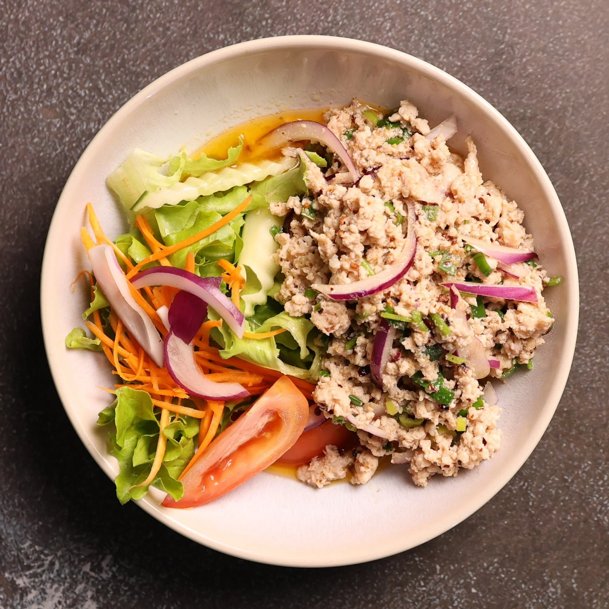 A bowl of Asian-inspired chicken salad with chopped vegetables including carrots, radish, lettuce, and cucumber, topped with ground chicken and a small serving of sauce or dressing.