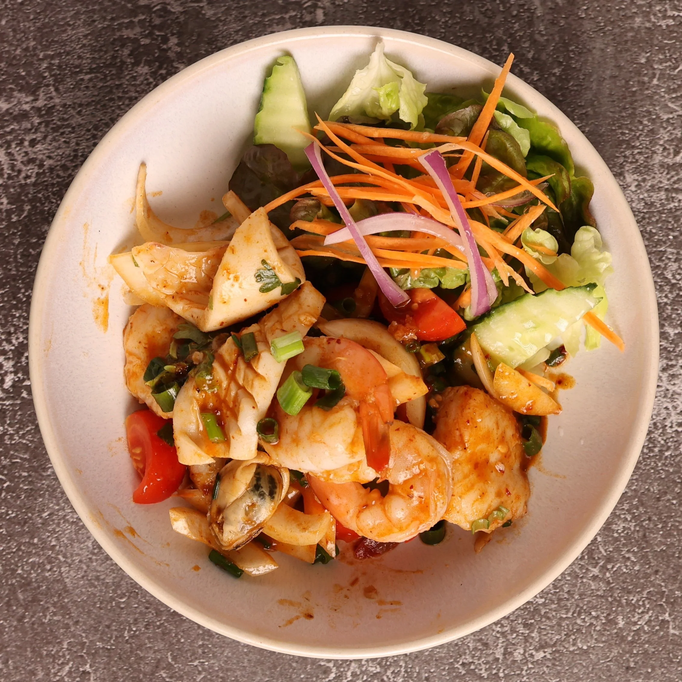 A white bowl filled with mixed vegetables, including shredded carrots, sliced cucumbers, lettuce, red onions, and pieces of cooked chicken, placed on a speckled dark gray countertop.