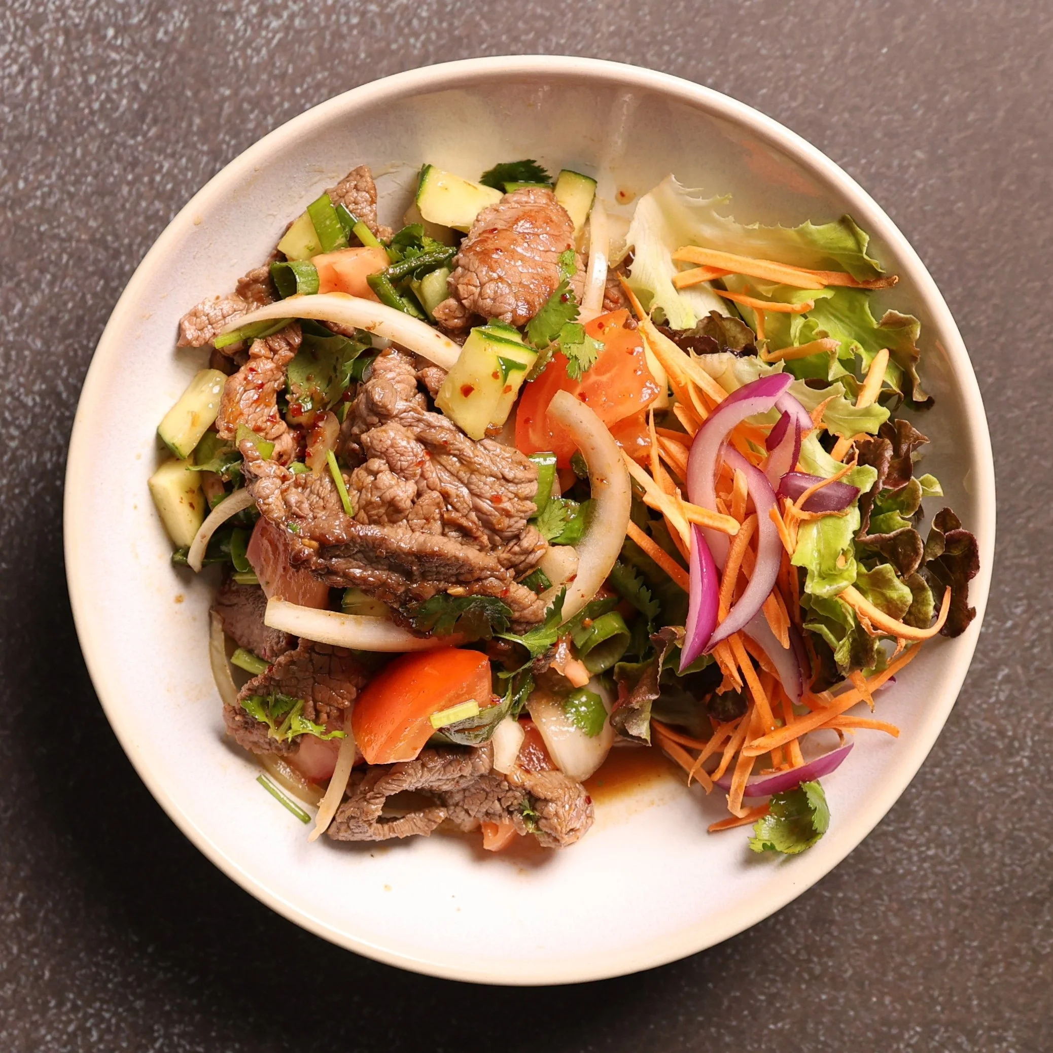 A white bowl filled with a salad containing sliced beef, chopped cucumbers, shredded carrots, sliced onions, chopped tomatoes, and leafy greens on a dark speckled countertop.