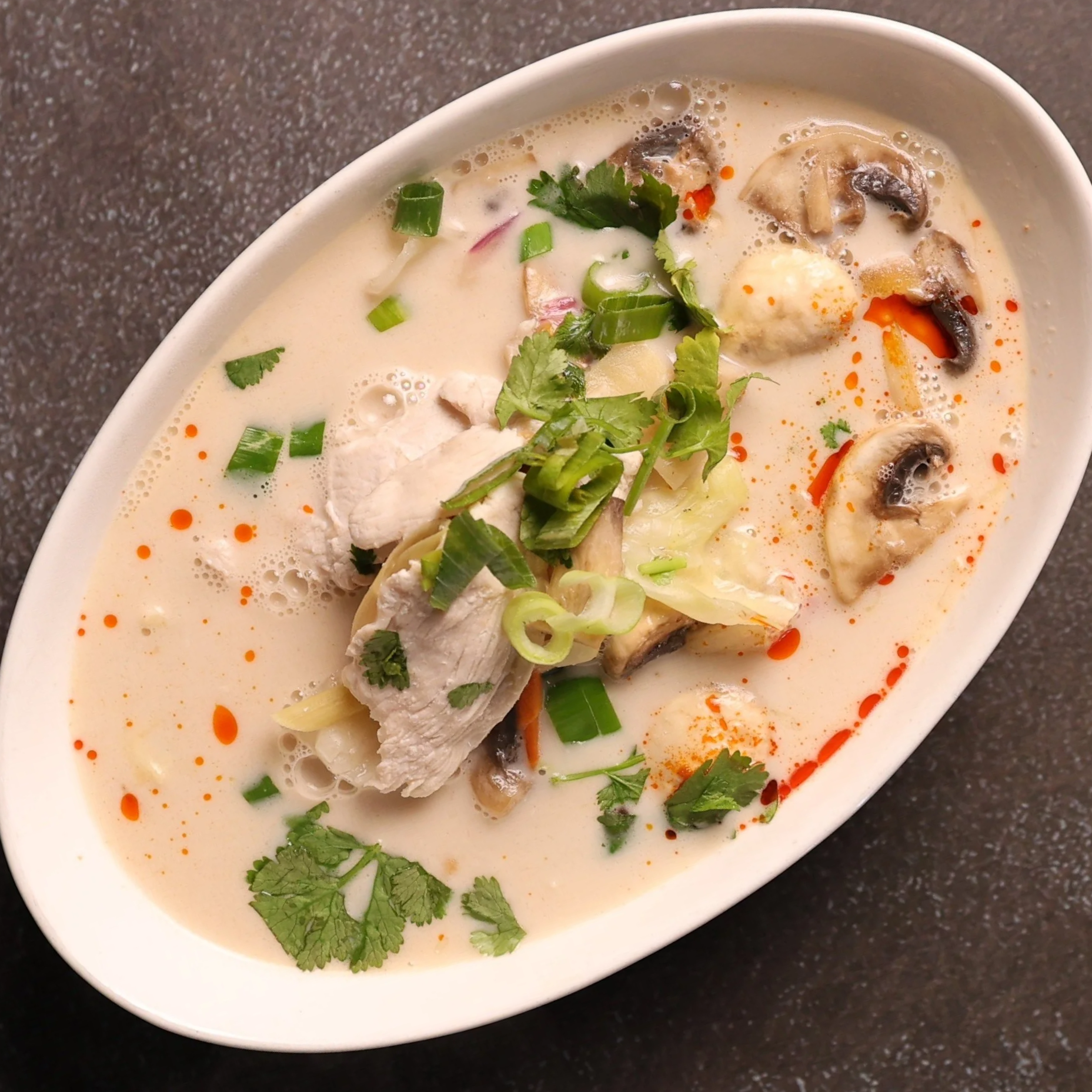 A bowl of Thai tom kha gai soup garnished with cilantro, green onions, red chili flakes, and cooked mushrooms.