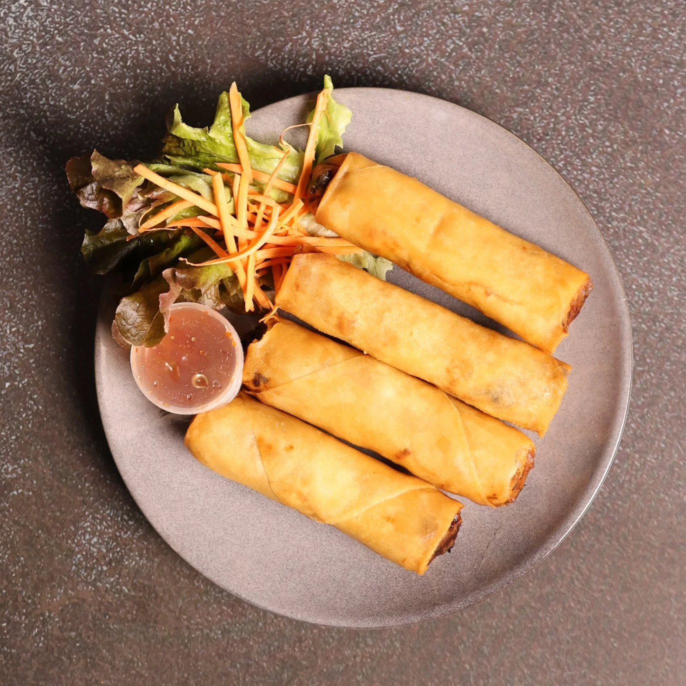 Four egg rolls on a plate with a side salad of lettuce, shredded carrots, and a small container of dipping sauce.