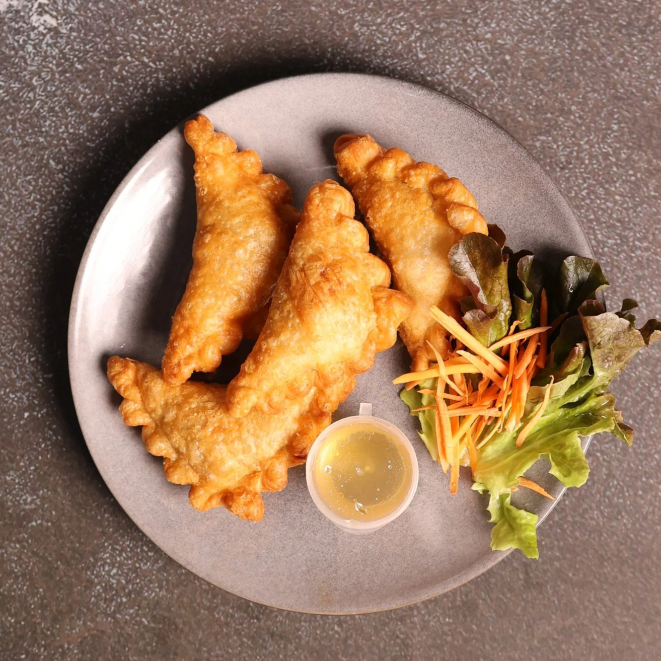 Fried fish fillets served on a gray plate with a small cup of tartar sauce and a side of mixed greens and shredded carrots.