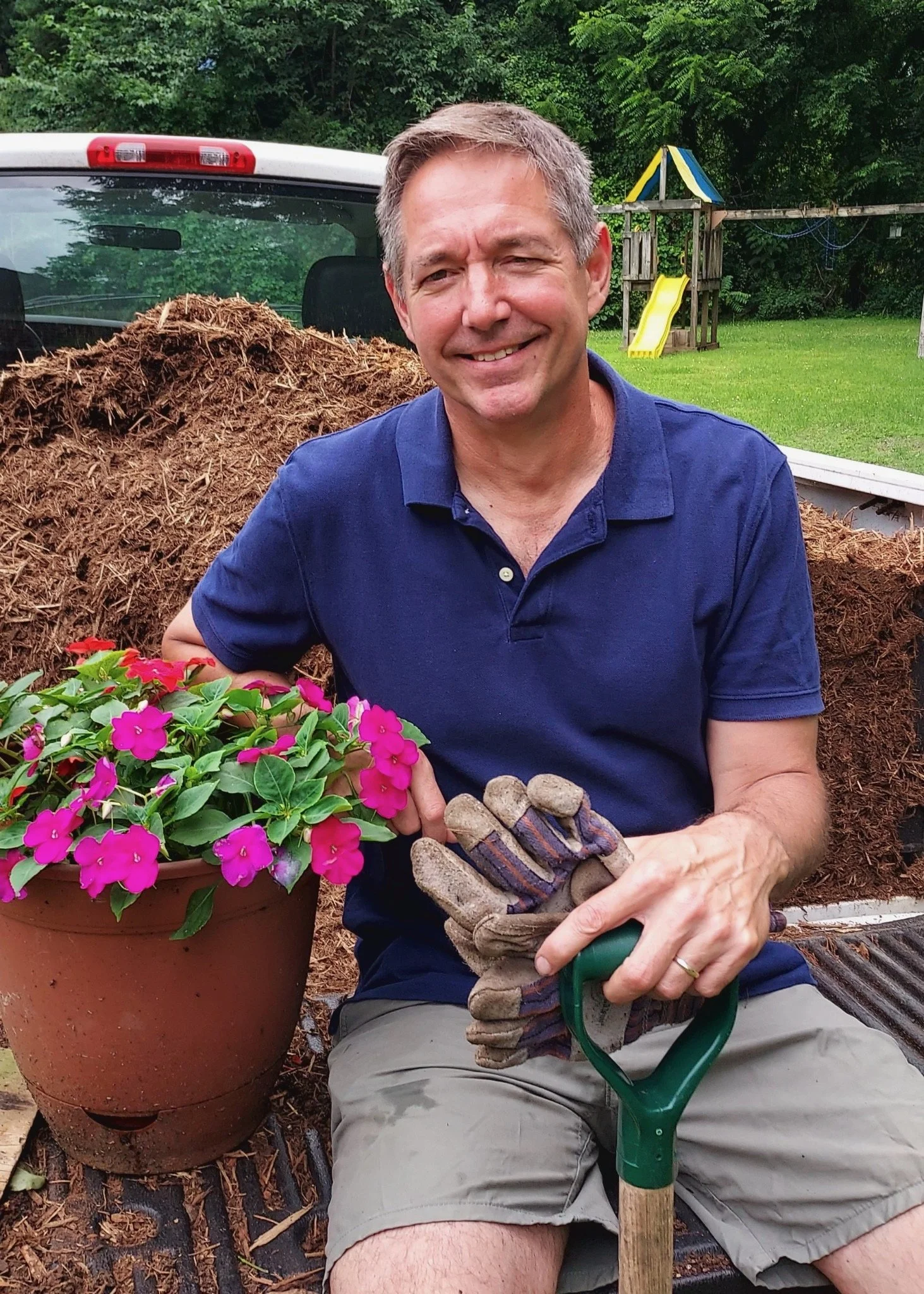 A middle-aged man sitting outdoors in a backyard, smiling, holding gardening gloves, surrounded by a potted pink flowering plant, a garden shovel, a pile of mulch, and a children's play structure in the background.